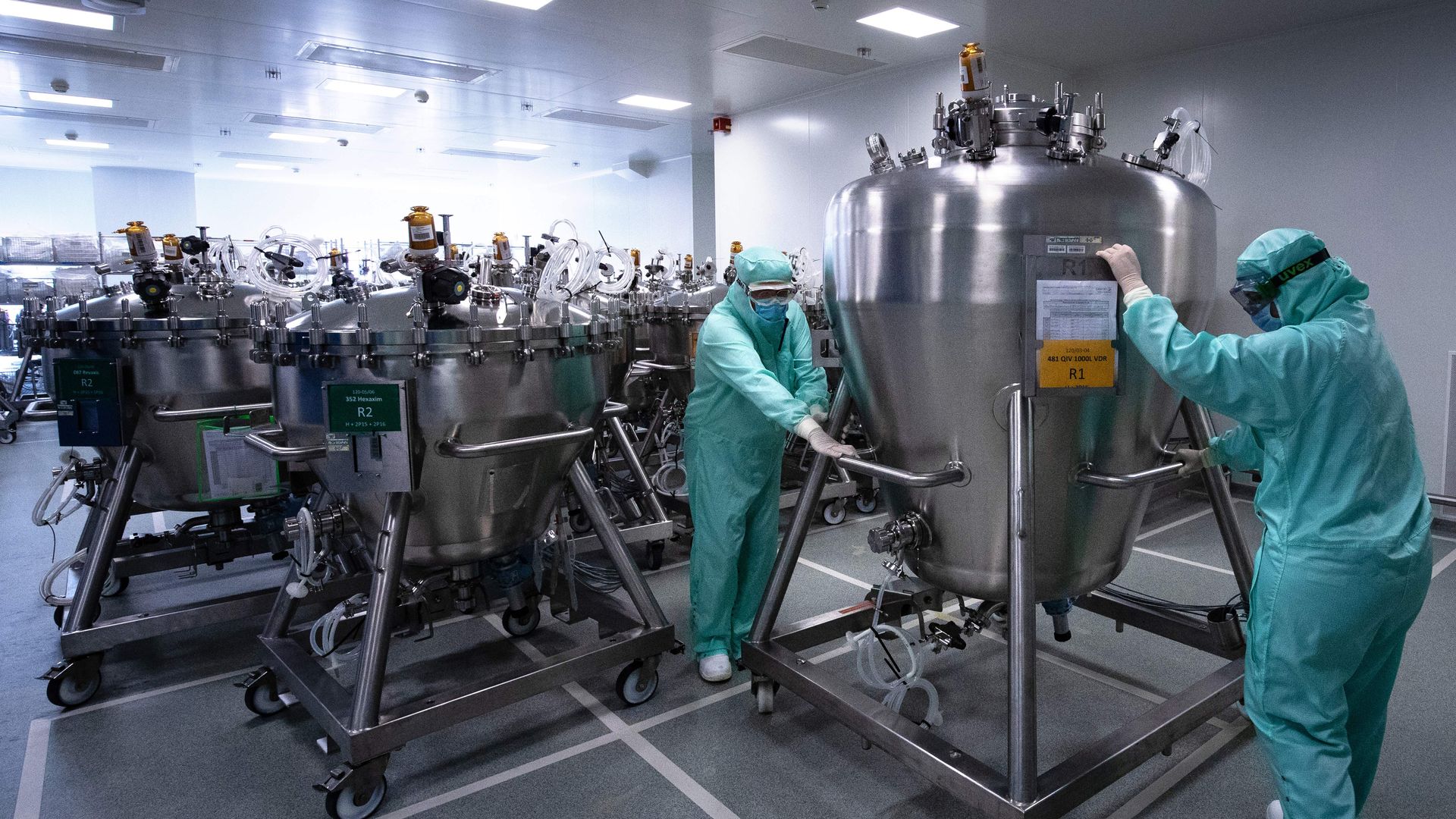 Two technicians in green protective equipment handle large pharmaceutical equipment.