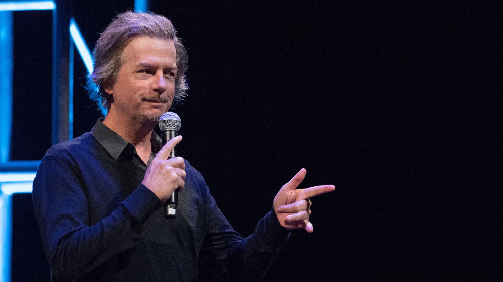 David Spade holding a microphone on stage and pointing