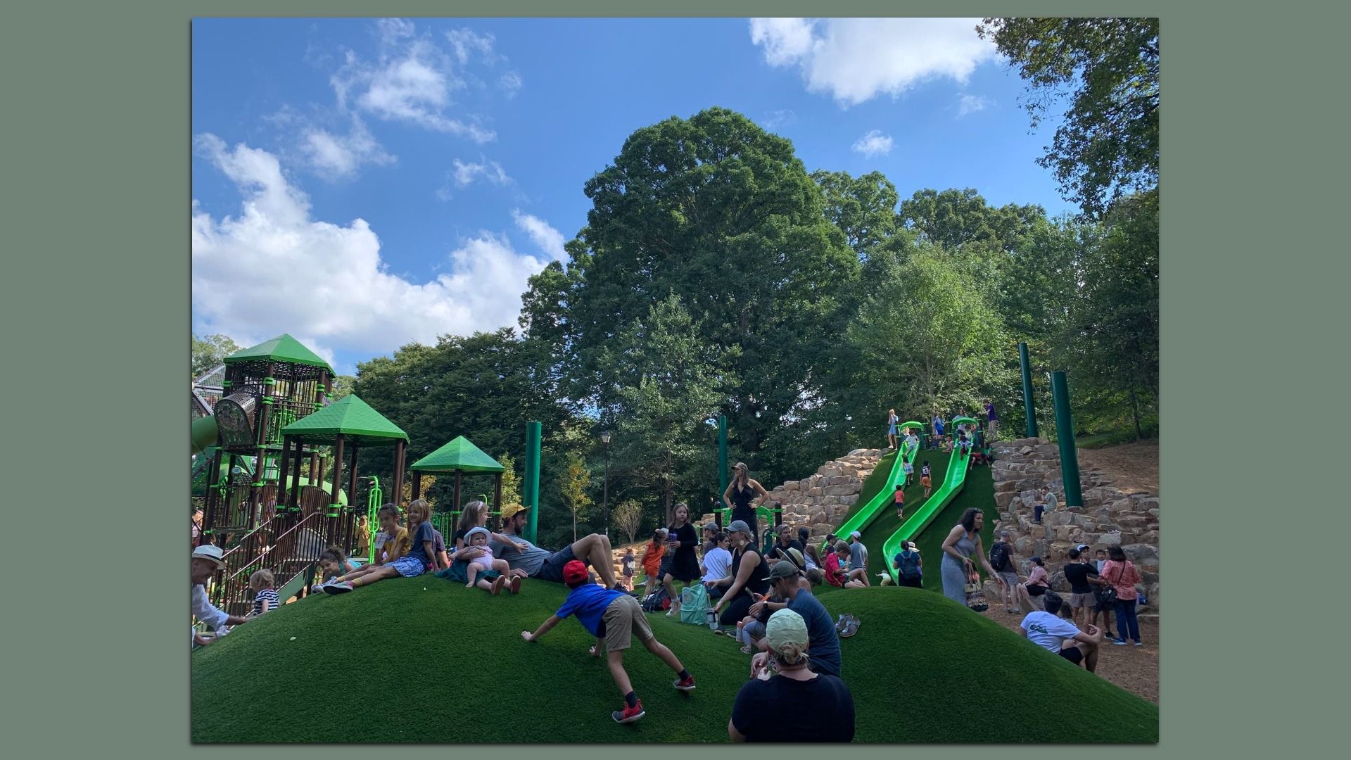 Playground with green slides and climbing structures surrounded by large trees under a blue sky with clouds. Many children and adults play and relax on grassy hills.