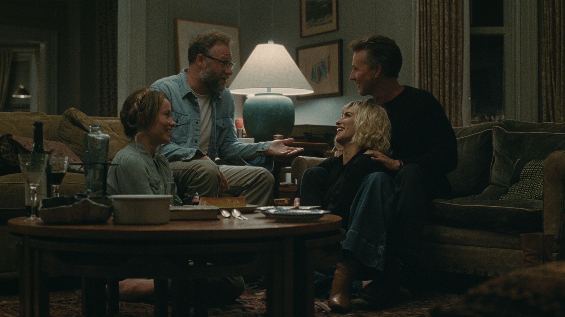 Four friends—two men and two women—sit around a coffee table in a dim living room, smiling and talking. A large blue lamp glows between them; drinks and snacks sit on the table.