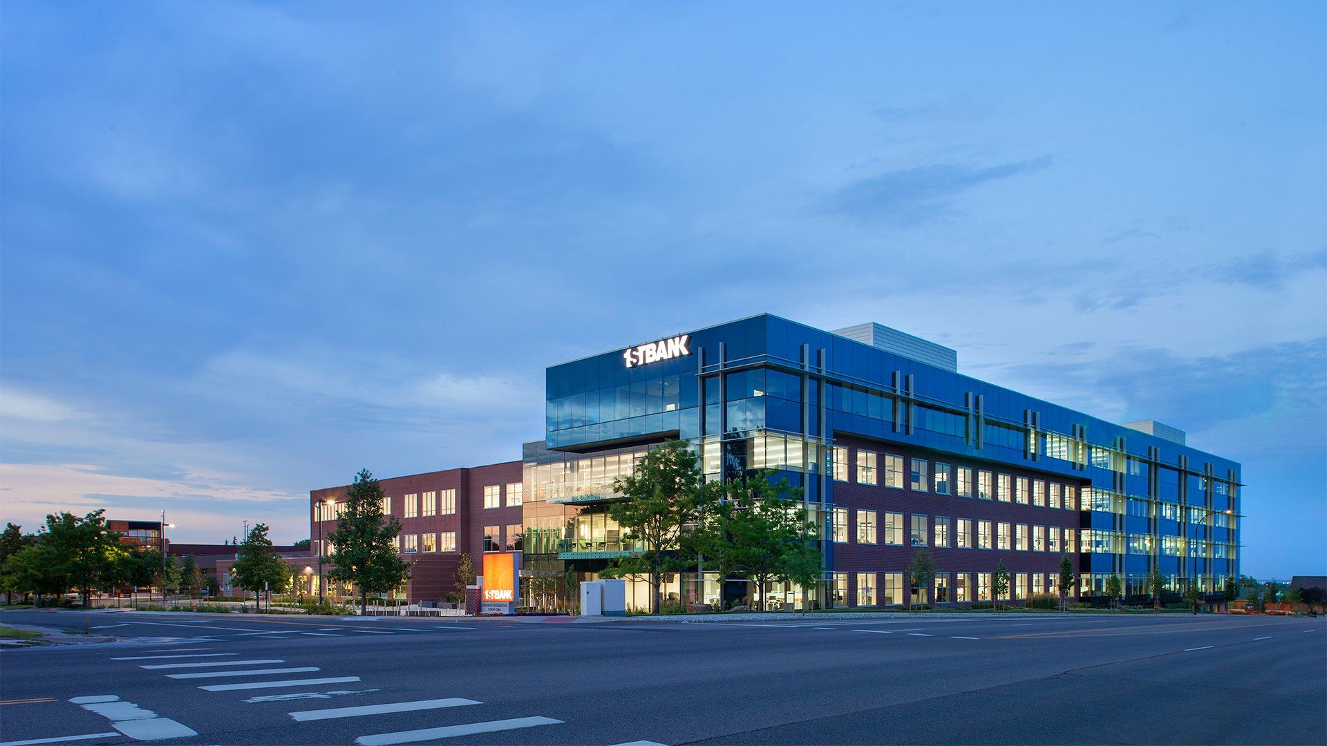 Modern office building with glass and brick exterior, labeled "1ST BANK," illuminated at dusk with a clear blue sky and empty street crosswalk in front.