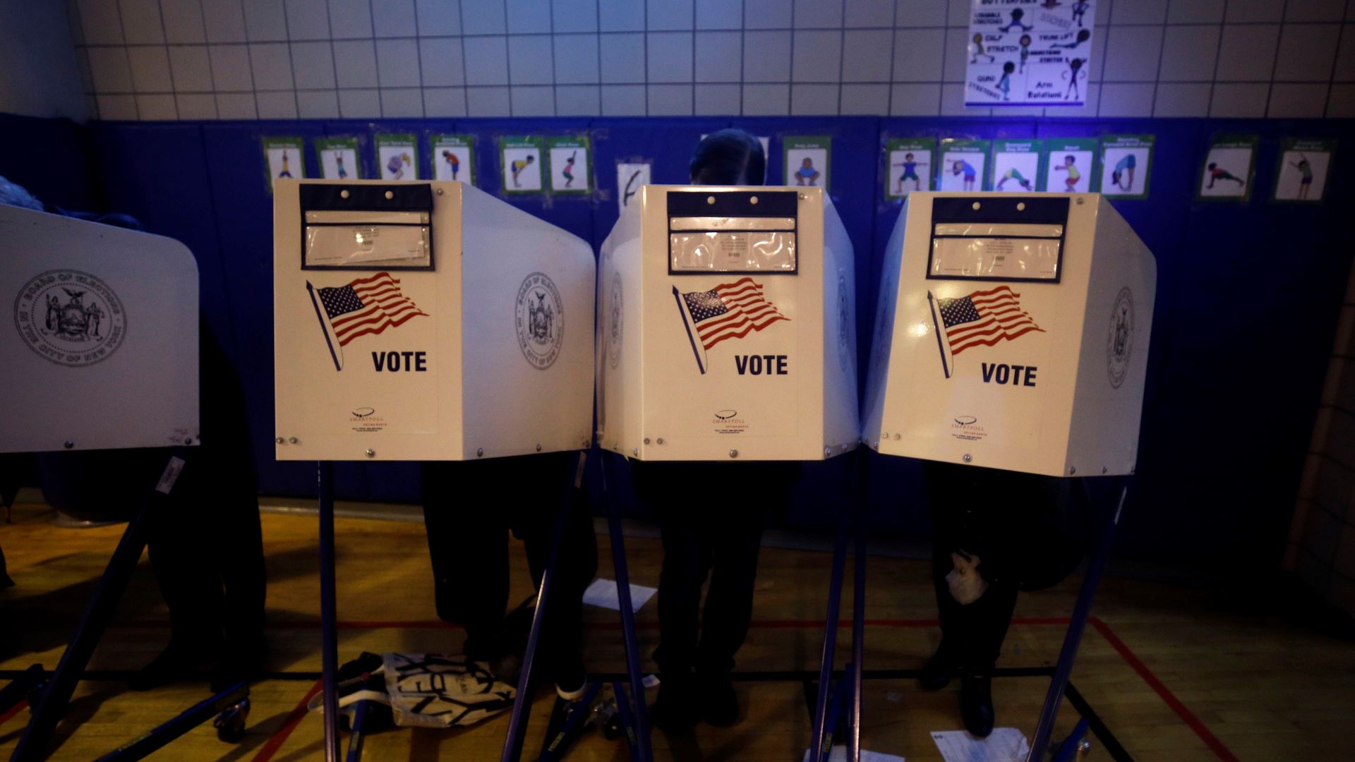 In this image, three people stand behind three different white voting booths. Each booth has an American flag on it.