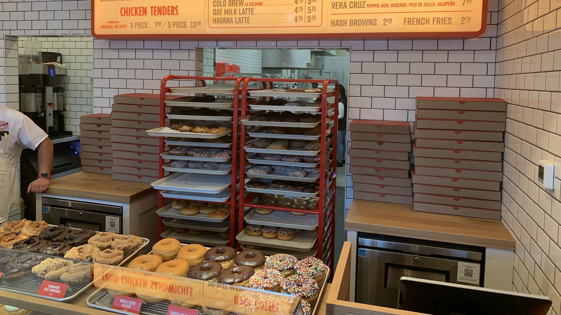 Inside a bright bakery: glass donut case, trays on a red rack, white subway-tile walls, and an orange menu board listing Fried Chicken Sandwiches, Drip Coffee, and Egg Sandwiches.