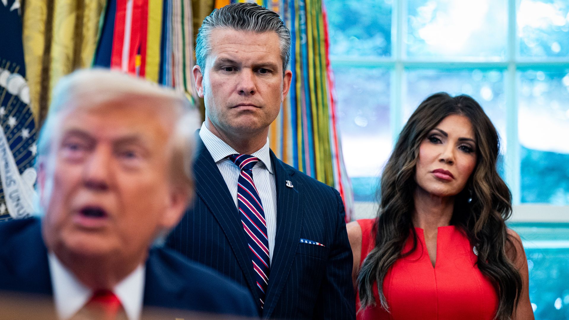 Donald Trump seated at a desk in the Oval Office signing documents as Pete Hegseth, the U.S. secretary of defense, and Kristi Noem, the U.S. secretary of homeland security, stand nearby during an executive order event.