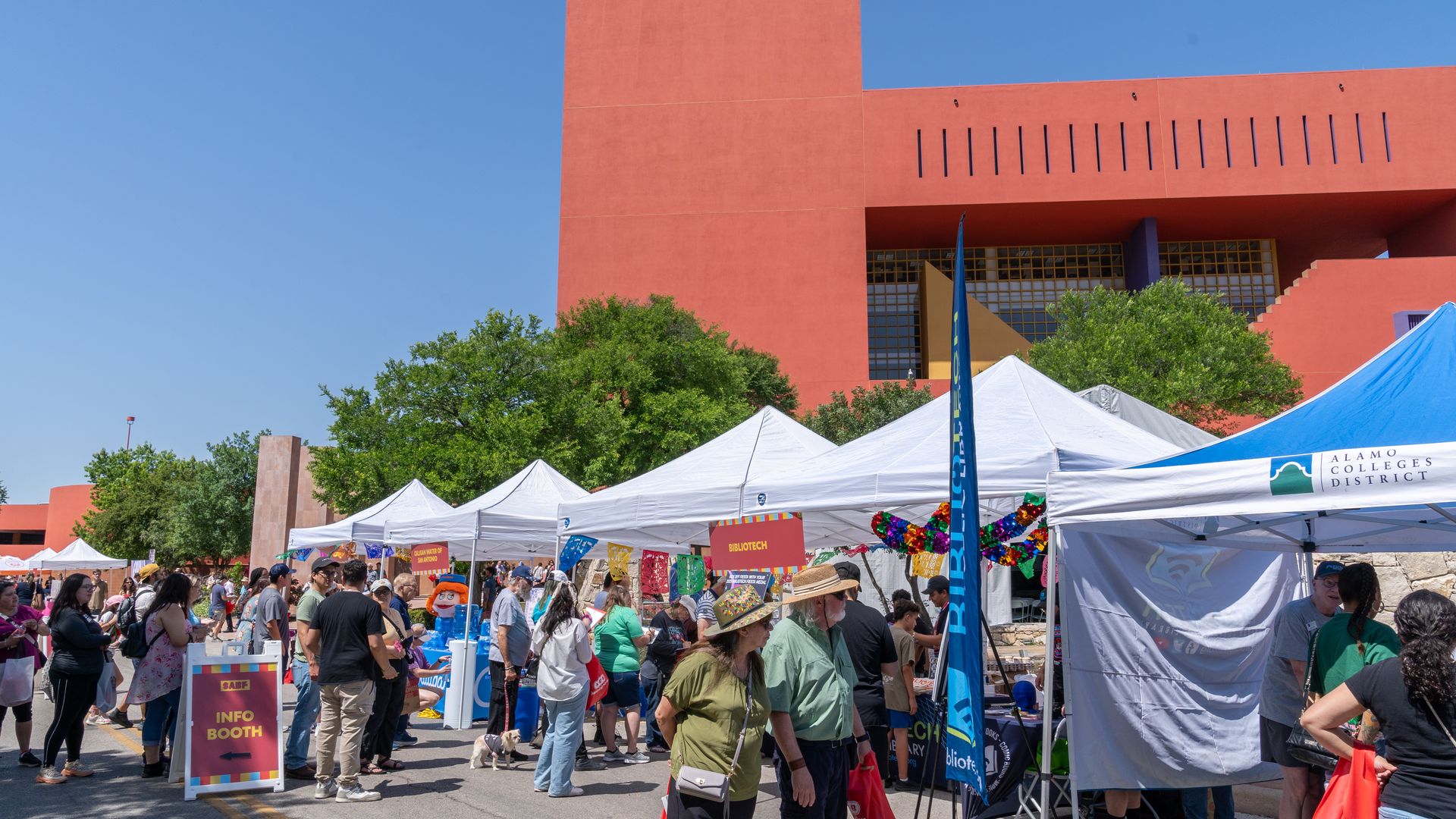 The San Antonio Book Festival as seen in 2025 with several white tents against the backdrop of a clear blue sky and a bright red Central Library. People are seen walking and browsing.