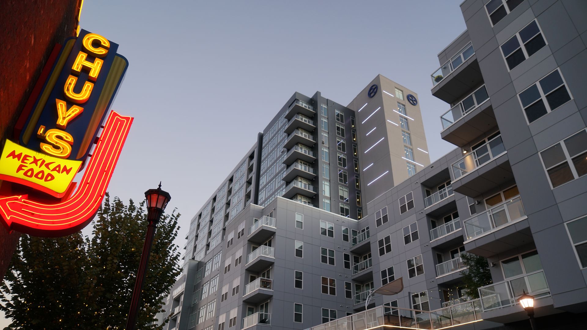 Exterior view of a modern gray apartment building at dusk with balconies and lights on. To the left is a bright neon sign for "CHUY'S Mexican Food" in yellow and red colors.