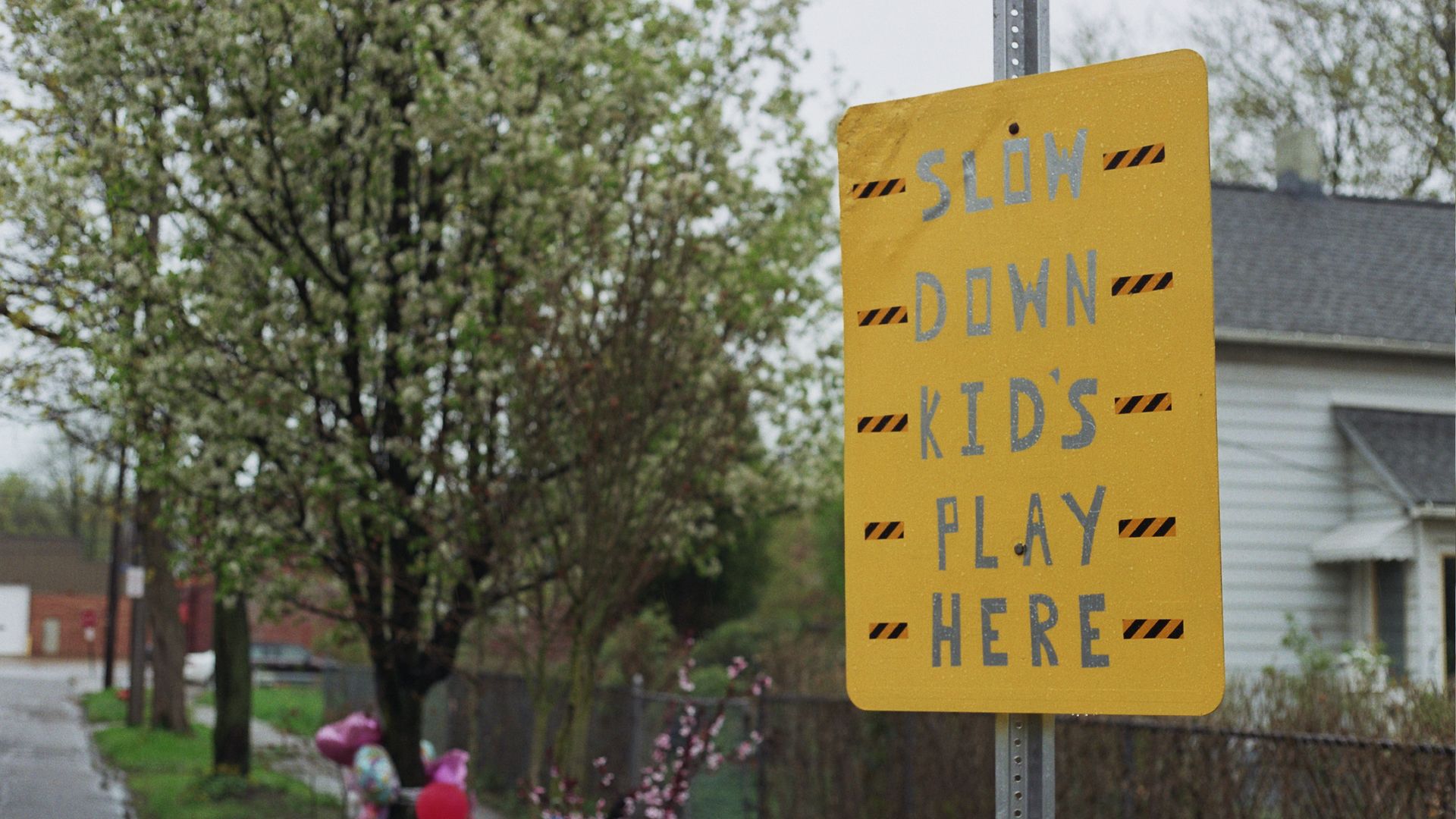 A handmade yellow sign reading, "Slow Down Kids Play Here" with a tree lawn memorial in the background