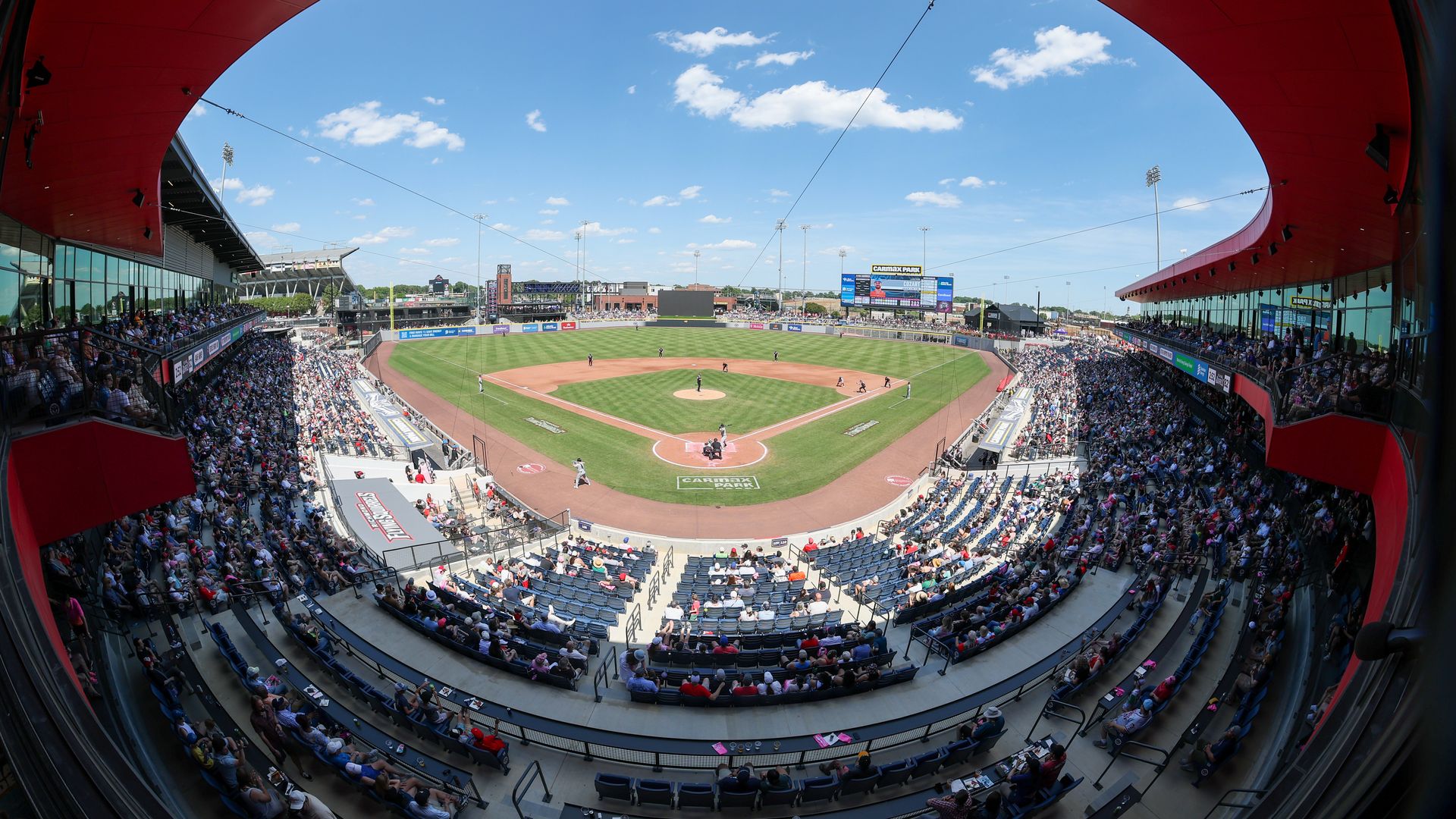 Panoramic view from the stands of a baseball stadium: a bright blue sky with fluffy clouds, a red curved roof framing the field, packed seats, and players on the green diamond with a distant scoreboard.