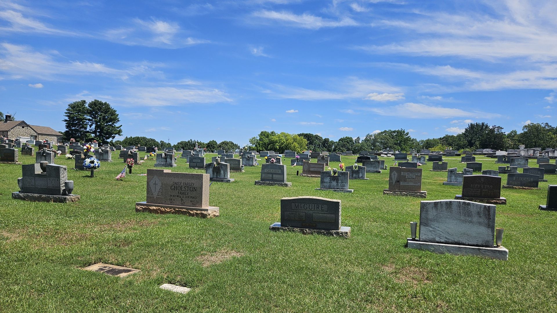 A sunny cemetery with rows of gravestones on green grass, a small stone building to the left, colorful flowers, and a bright blue sky with wispy white clouds.