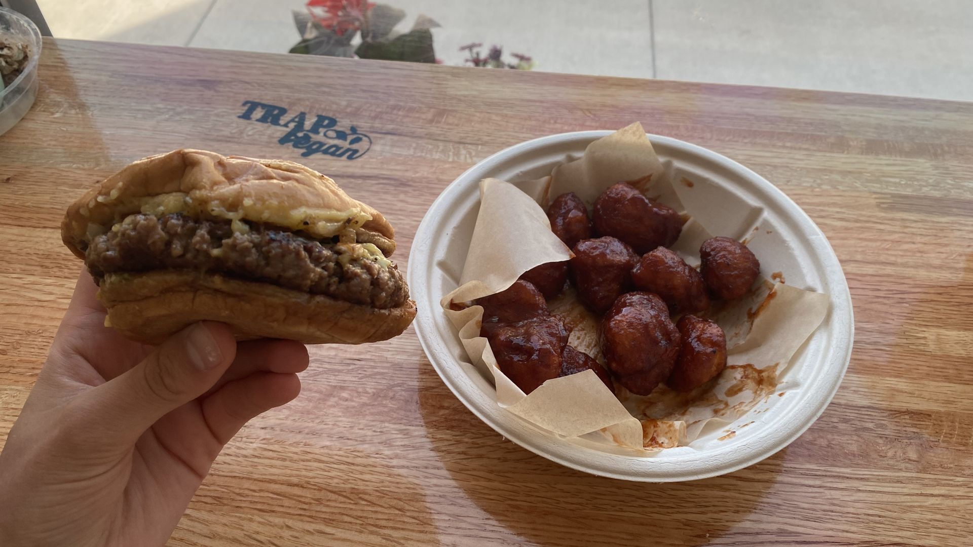 A burger, held in a hand, is shown next to a bowl of cauliflower wings on a table.