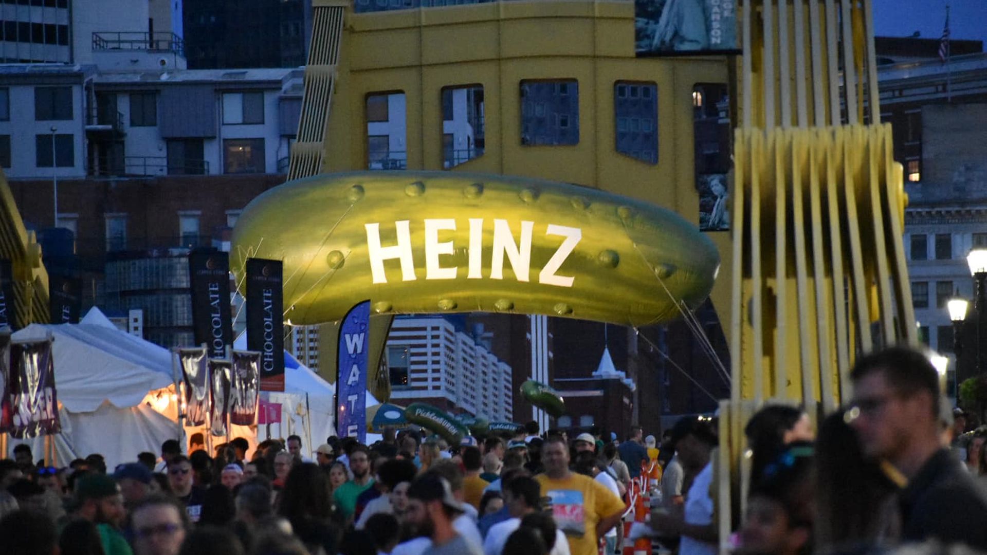At dusk in a busy city, a large yellow Heinz inflatable arch spans the street at a festival, flanked by white tents and banners as a crowd walks beneath.