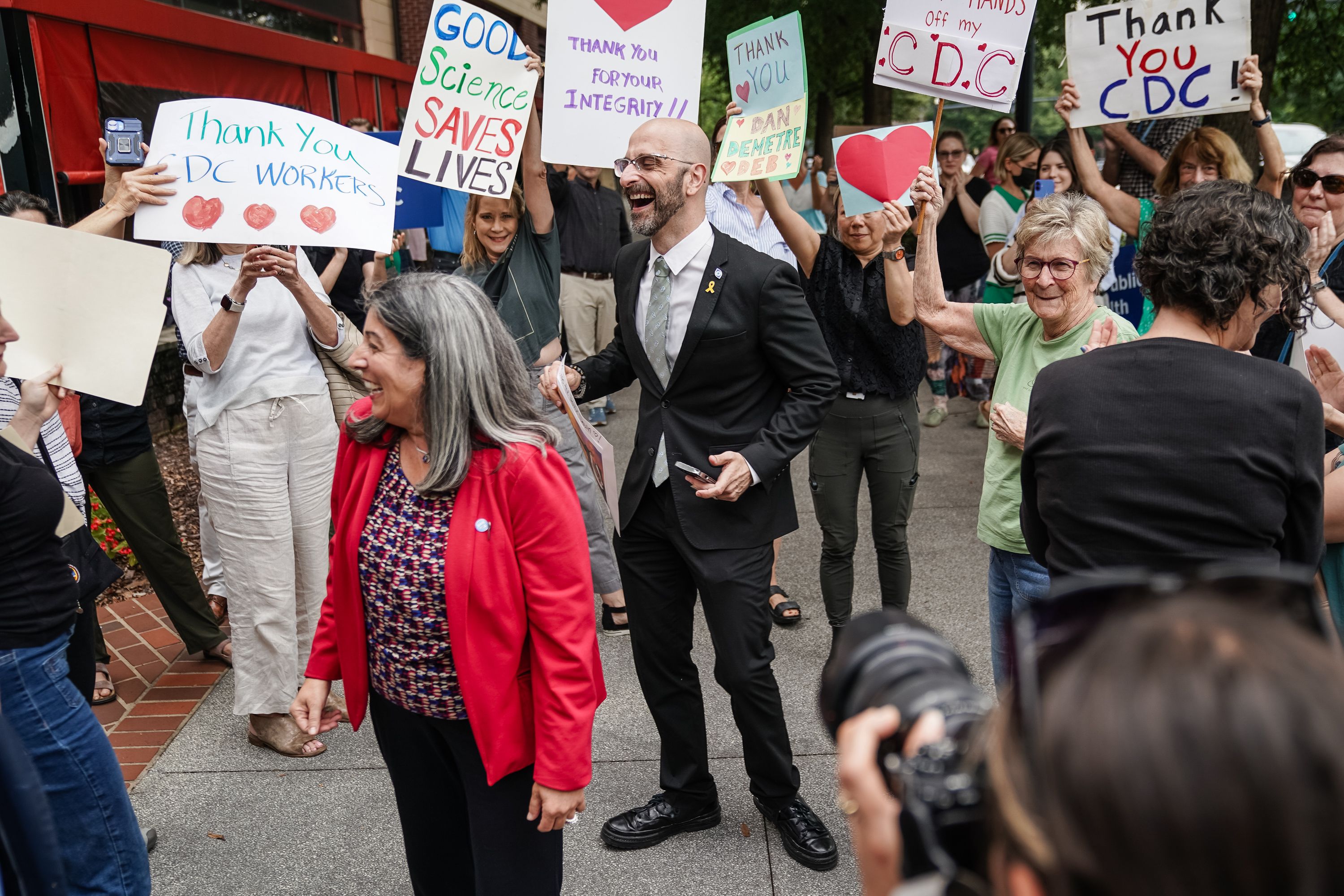 Group of diverse people outdoors holding signs thanking CDC and DC workers, with messages like "Good Science Saves Lives" and "Thank You CDC," smiling and clapping on a city sidewalk.