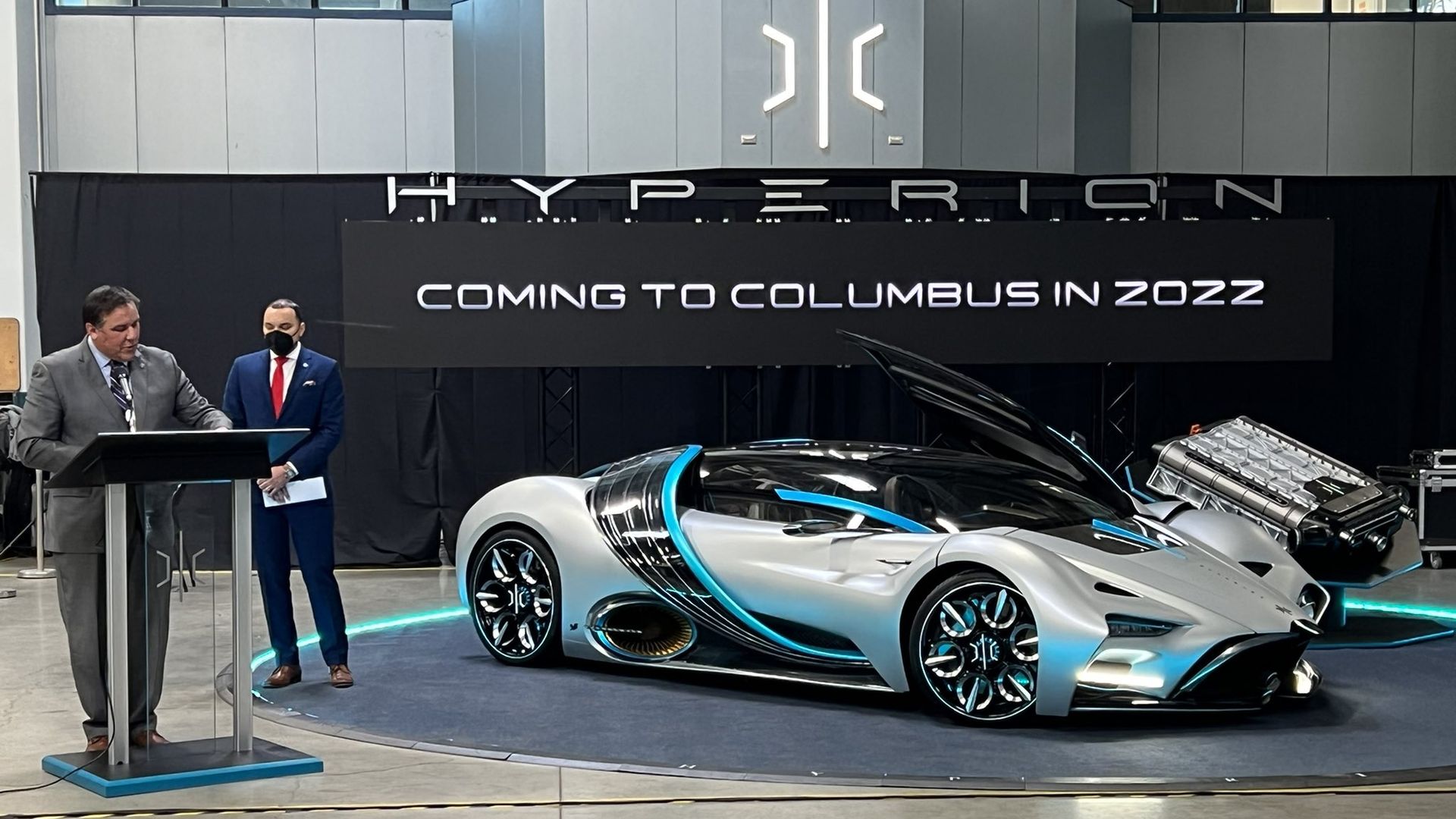 The Columbus mayor speaks at a podium next to a supercar with a banner reading "Coming to Columbus in 2022" in the background.