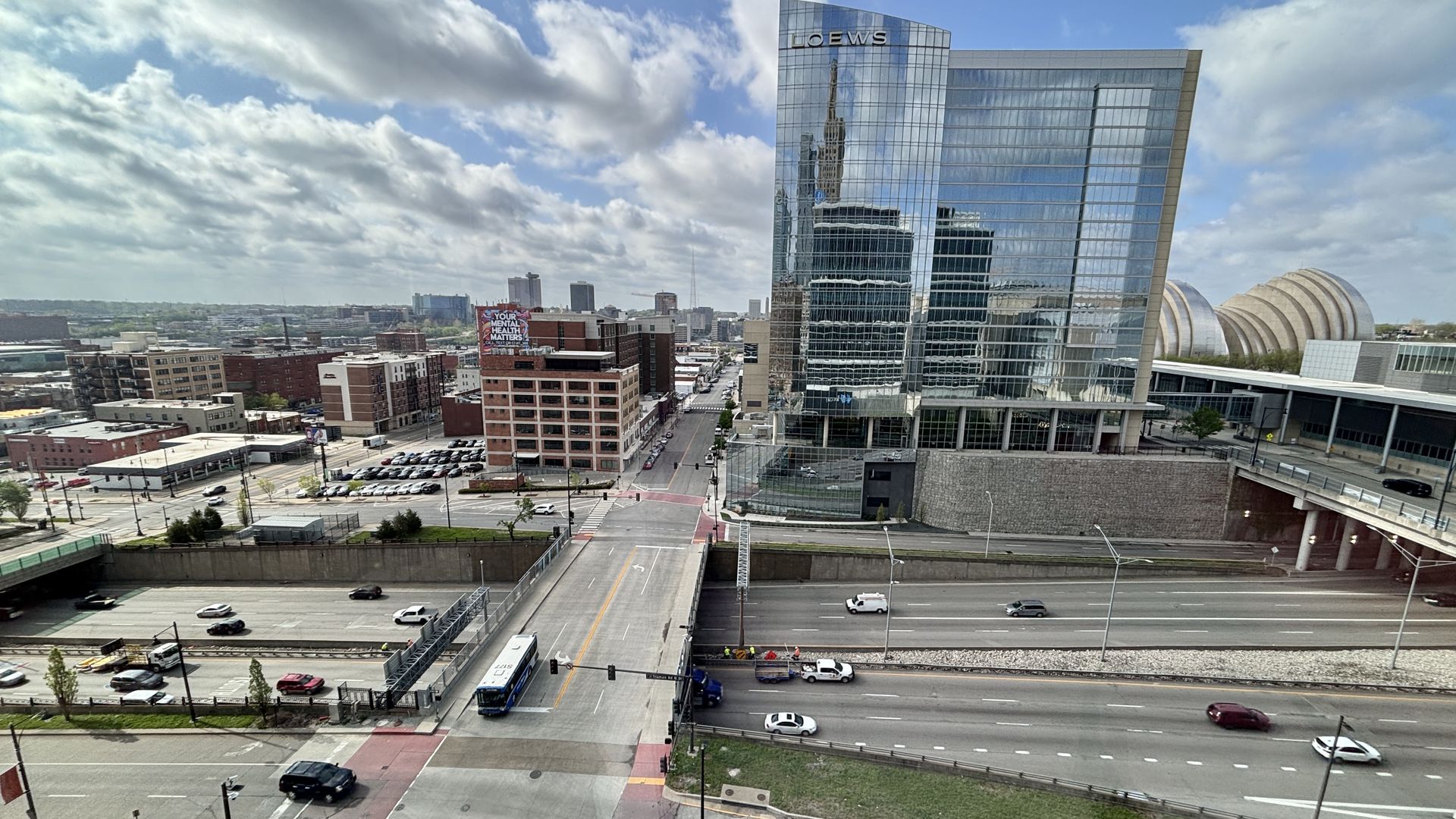An aerial city view: a tall glass Loews building on the right reflects the skyline; a busy road with a blue bus below, brick buildings, and a distant billboard.