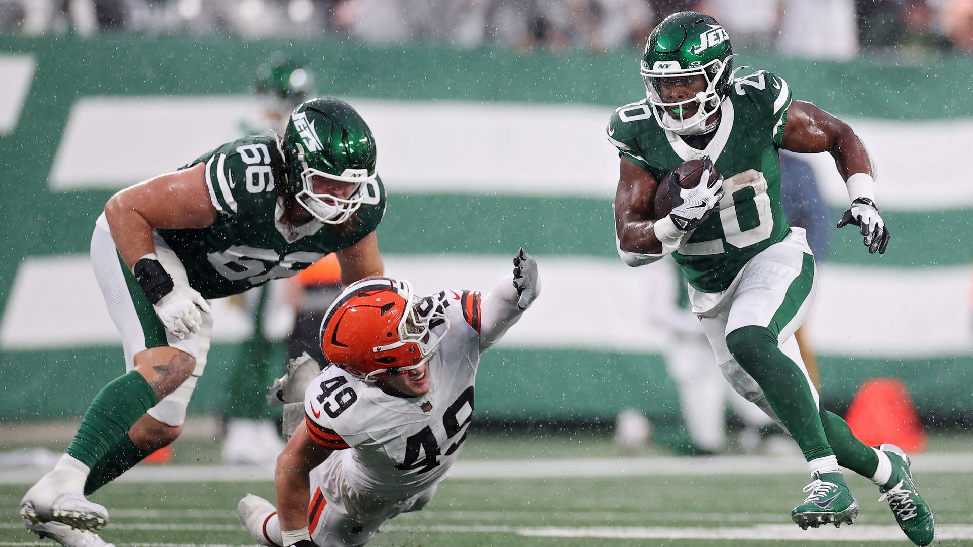 Football game in rain: player 20 in green Jets uniform runs with ball, player 49 in white Browns uniform falls trying to tackle, player 66 in green blocks opponent on field.