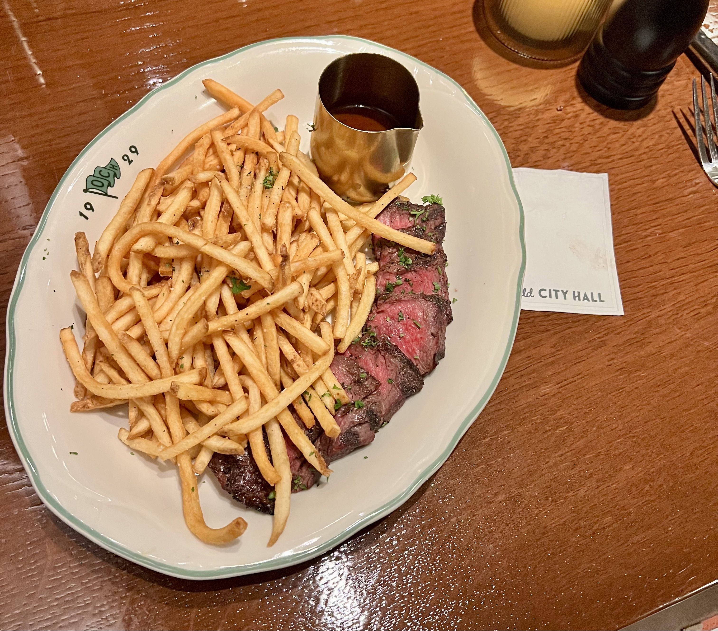 Plate of steak frites: slices of medium-rare steak with herbs, a large pile of thin French fries, and a small metal pitcher of sauce on a white plate with a green rim.