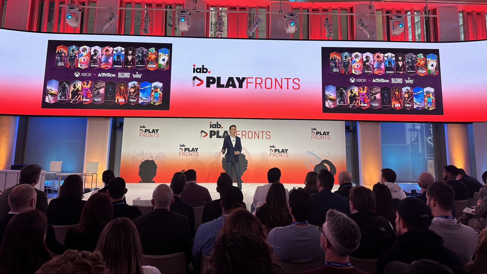 A man in a suit stands on a stage. A screen behind him says IAB PlayFronts and has a slide about Activision Blizzard's popular games. A seated crowd is in front of him
