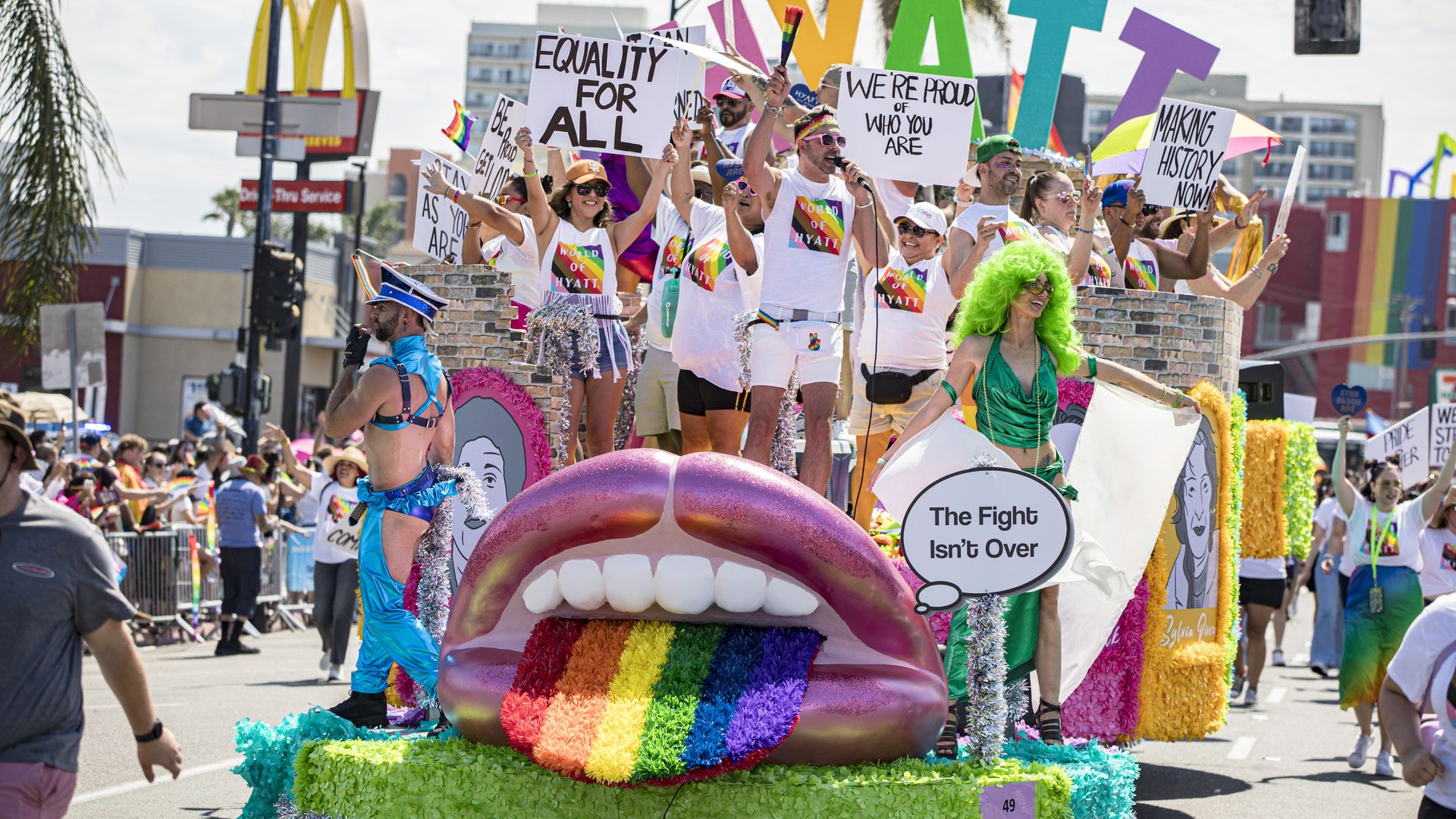 A Pride float with a large mouth and a rainbow tongue, plus dancers and people holding signs standing on it during a parade. 