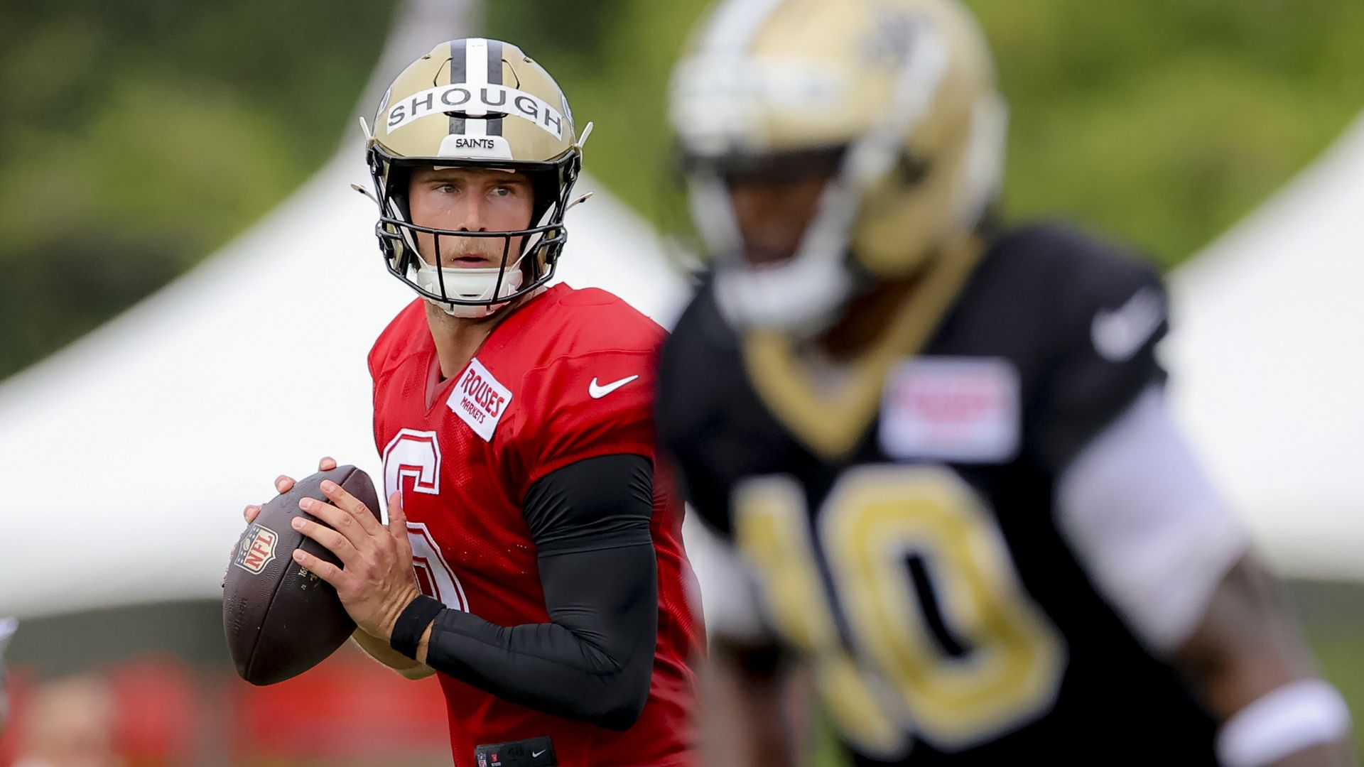 Football player in a red jersey and gold helmet labeled "SHOUGH" holding a football, preparing to throw, with another player in a black jersey and gold helmet blurred in the foreground.