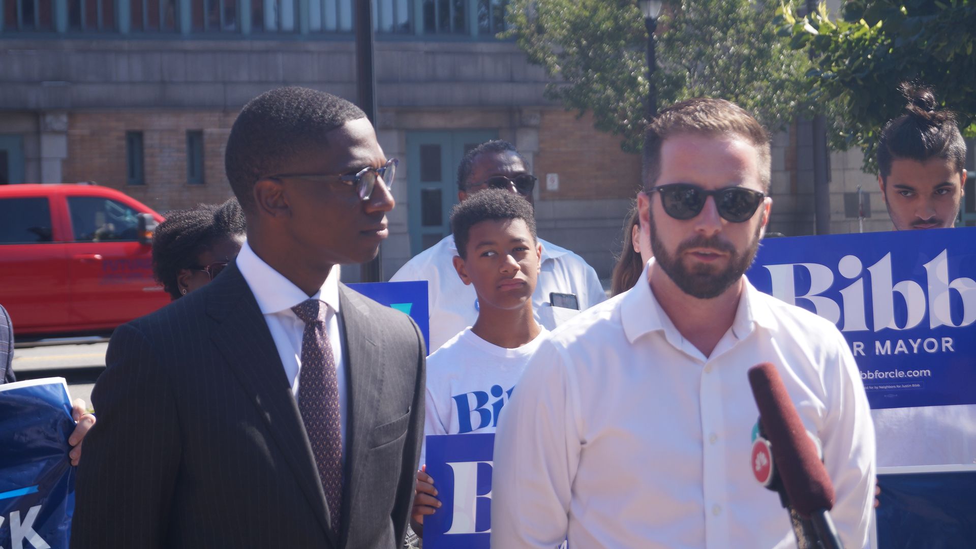 A group of people at an outdoor event, including a man in a white shirt with sunglasses speaking into a microphone, a man in a suit and tie, and others holding blue campaign signs reading "Bibb for Mayor".