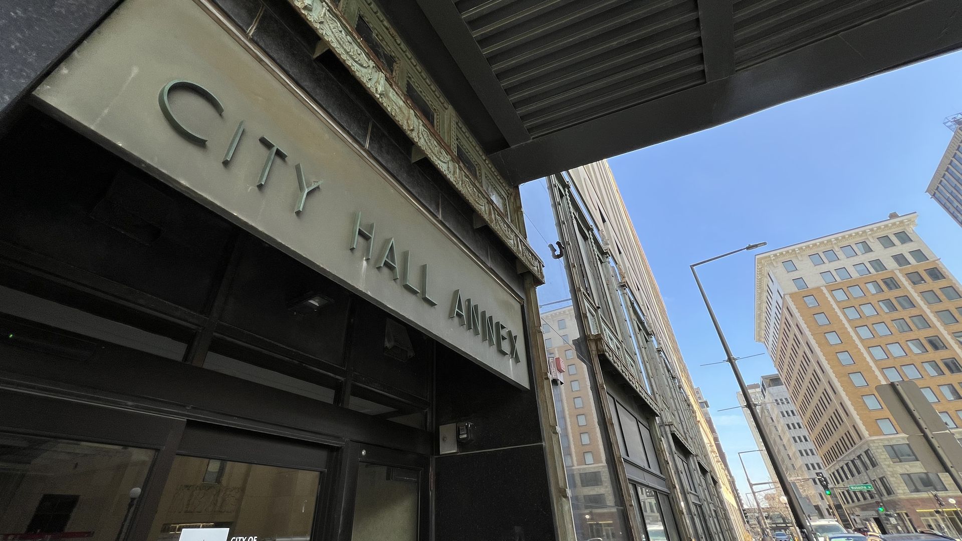 A sign on the exterior of a building in faded art deco style says "City Hall Annex." A downtown skyline stretches upward in the background.