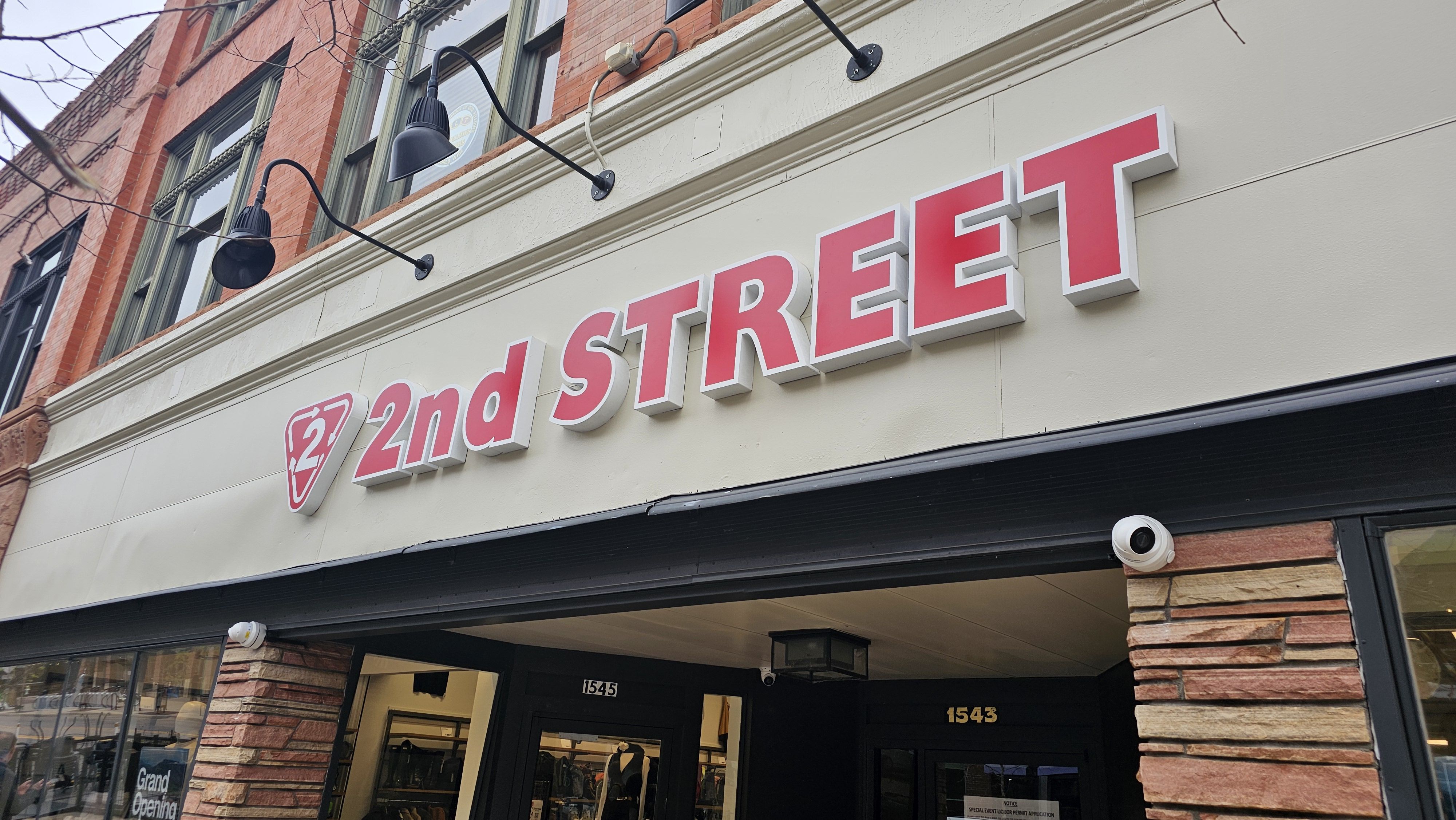 Storefront with a large pink 2nd Street sign on a beige wall, brick accents, and black gooseneck lamps. Glass windows; door numbers 1545 and 1543; Grand Opening sign visible.