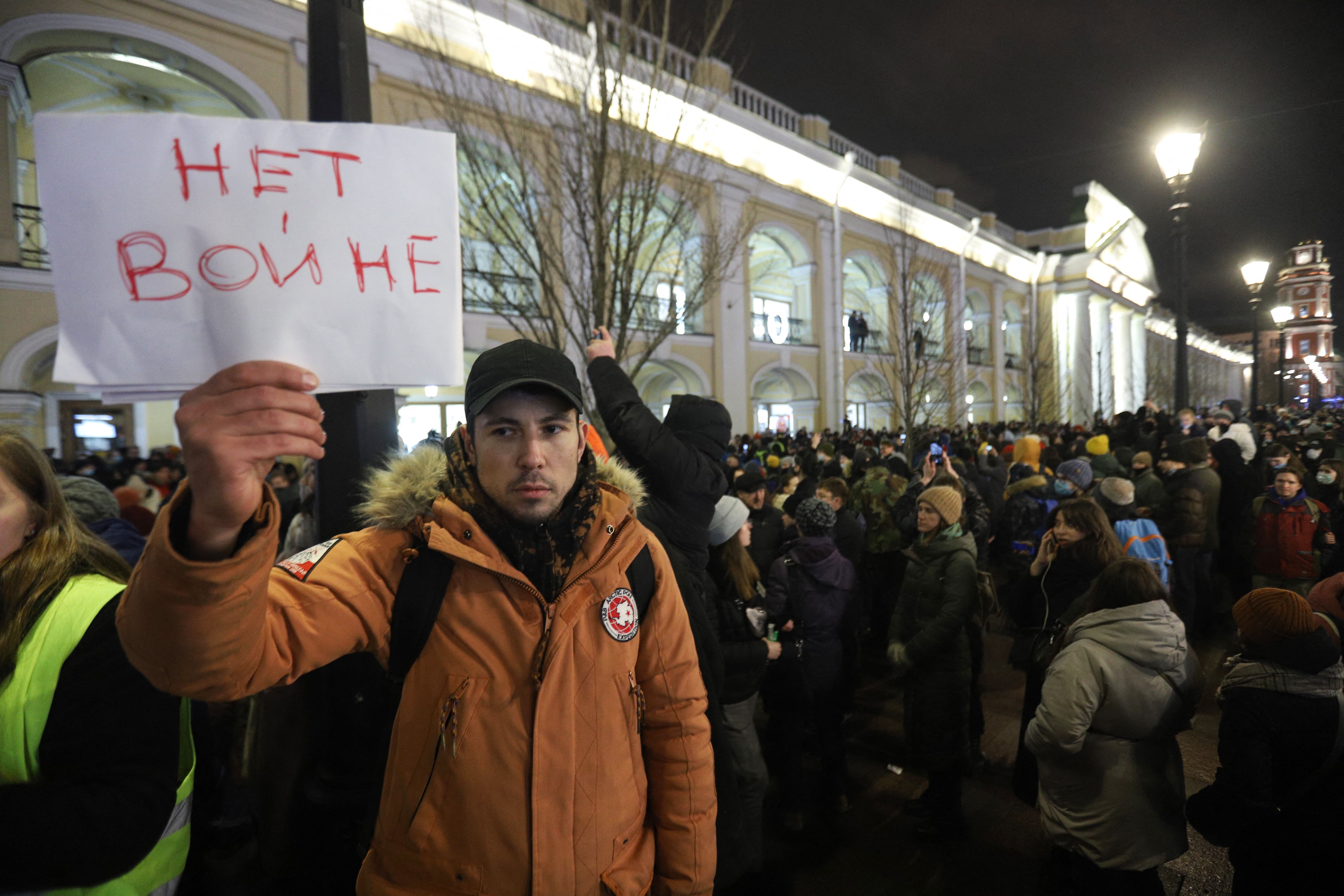 Photo of a person holding a sign that says "No to war" in Russian as they stand with a crowd of protesters