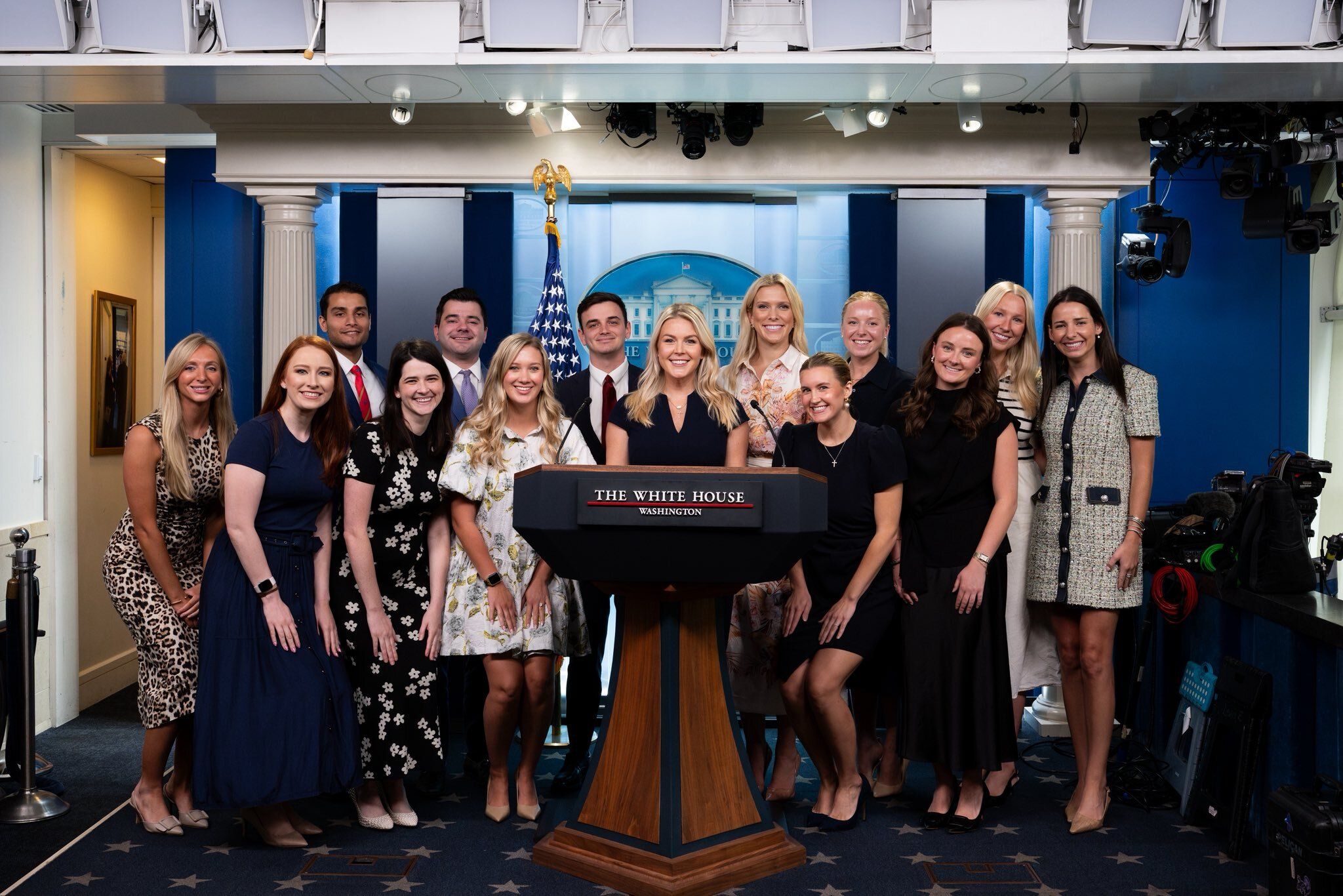 Group of men and women posing behind a podium in the White House briefing room, with the American flag, blue backdrop, and studio lights overhead.
