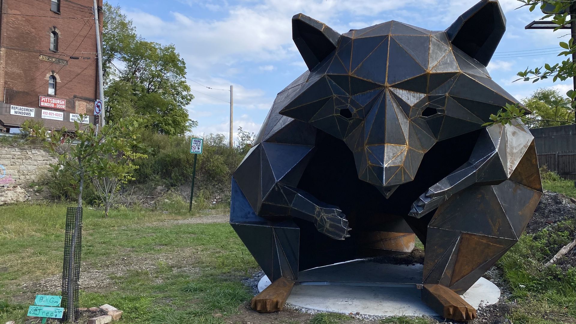Large geometric metal sculpture of a raccoon with angular shapes, set outdoors on grass near a brick building and small tree with a protective cage.