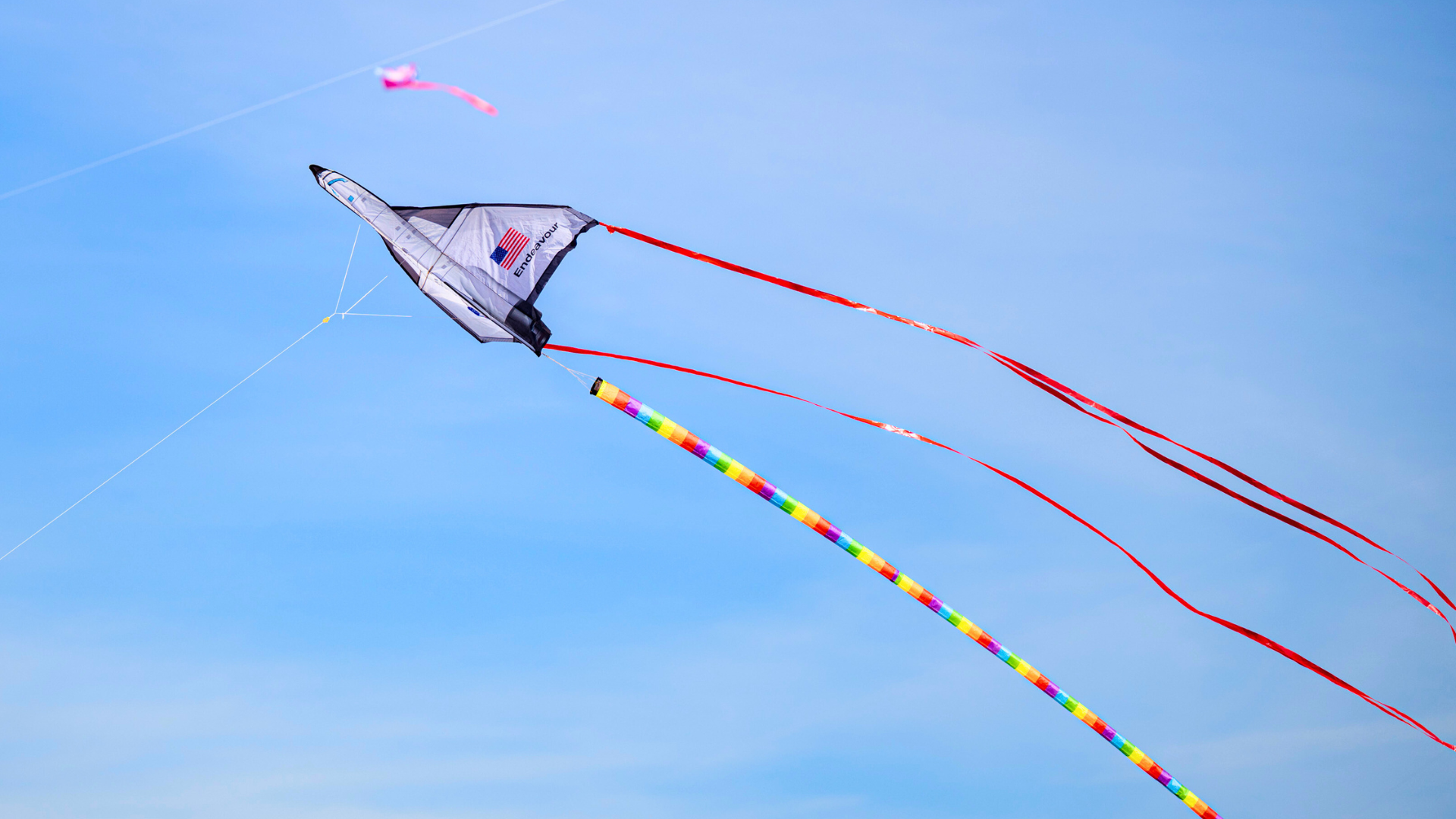 Gray and black delta kite with American flag and the word "Endeavour" flying in a clear blue sky, trailing long red tails and a rainbow-colored tail below.