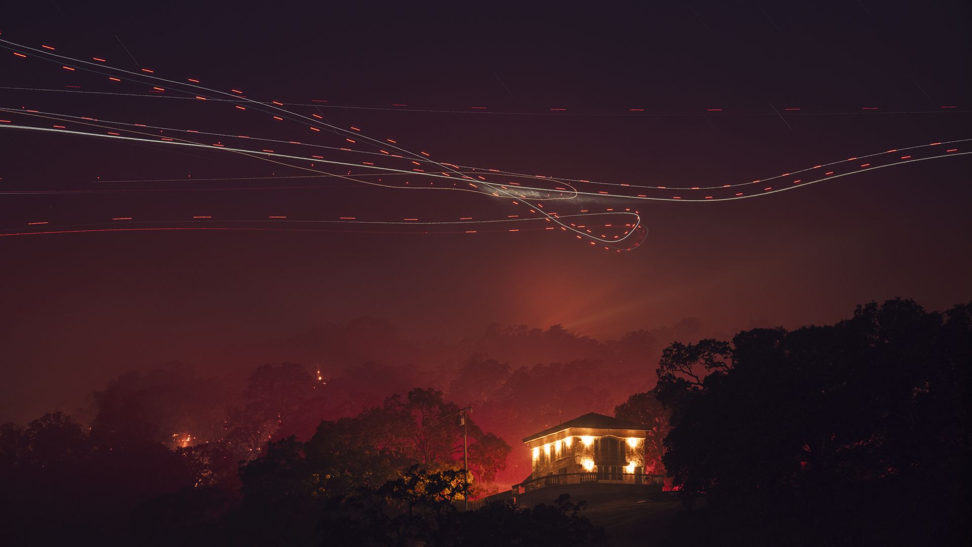 Long-exposure night scene: purple sky with star trails and red/white light trails; a warmly lit house sits on a hill among dark trees, with an orange glow at the horizon.