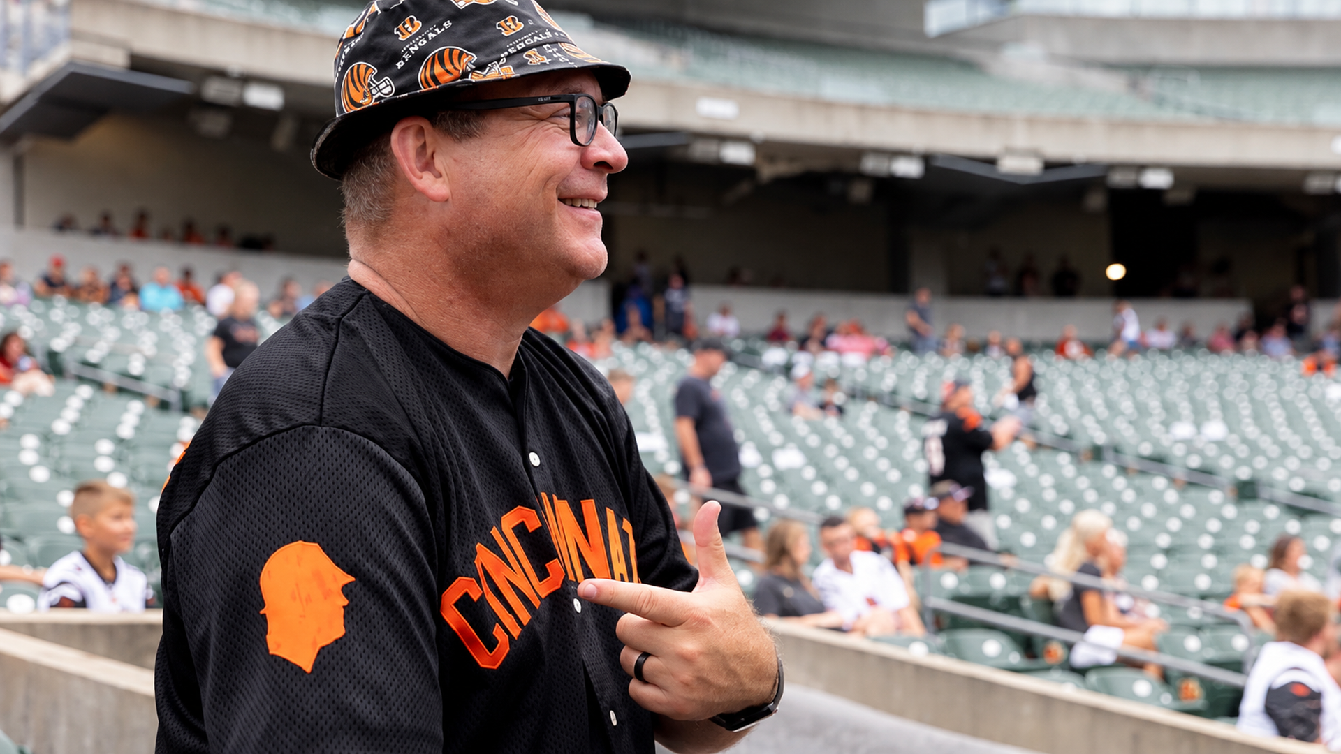 Smiling man in a black Cincinnati jersey with orange lettering and a tiger-pattern hat, stands in a stadium with green seats and blurred fans in the background.