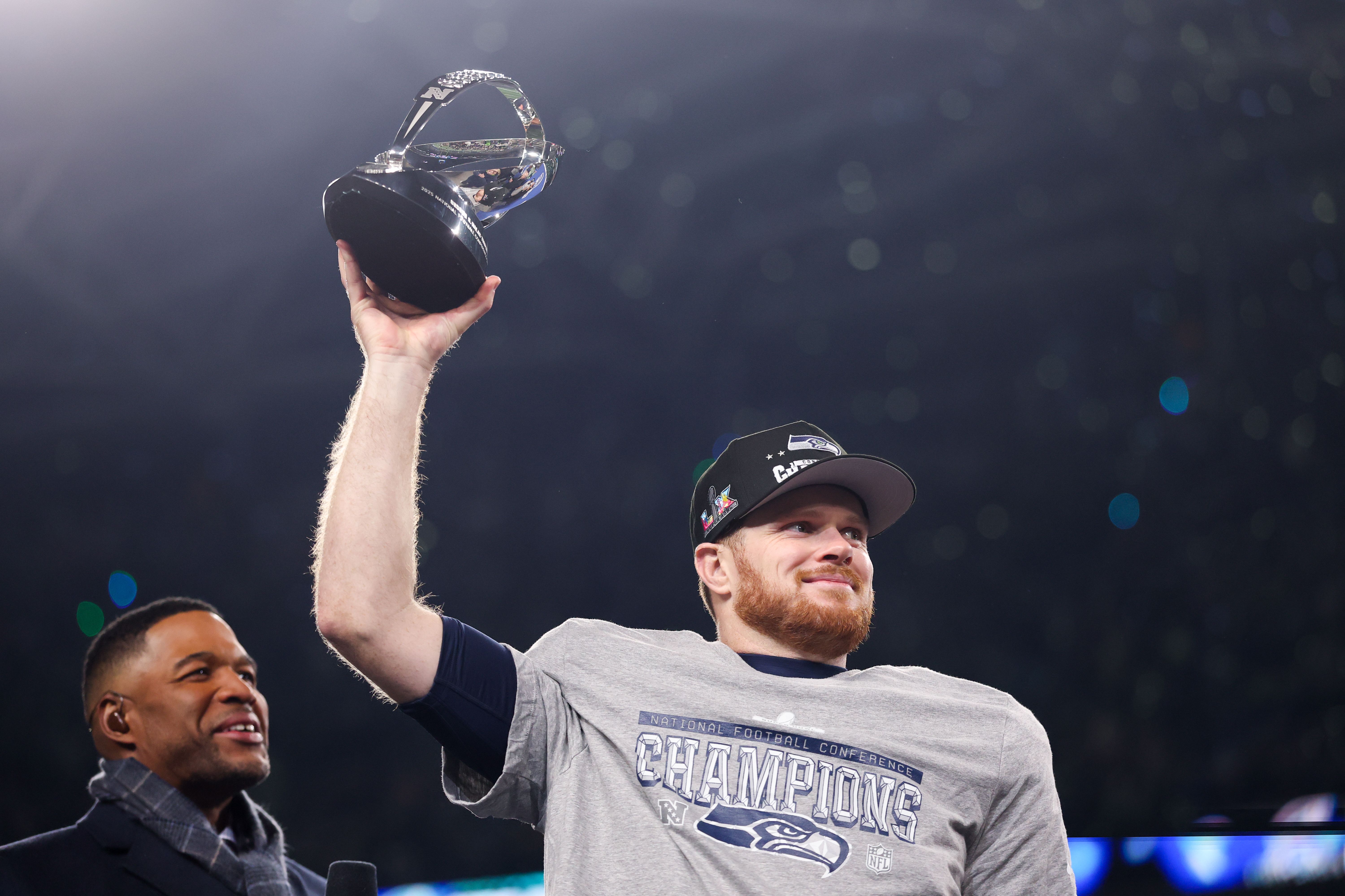 Seahawks quarterback Sam Darnold wears a ball cap and a gray T-shirt that says National Football Conference Champions, while holding the NFC championship trophy over his head in  the stadium. 