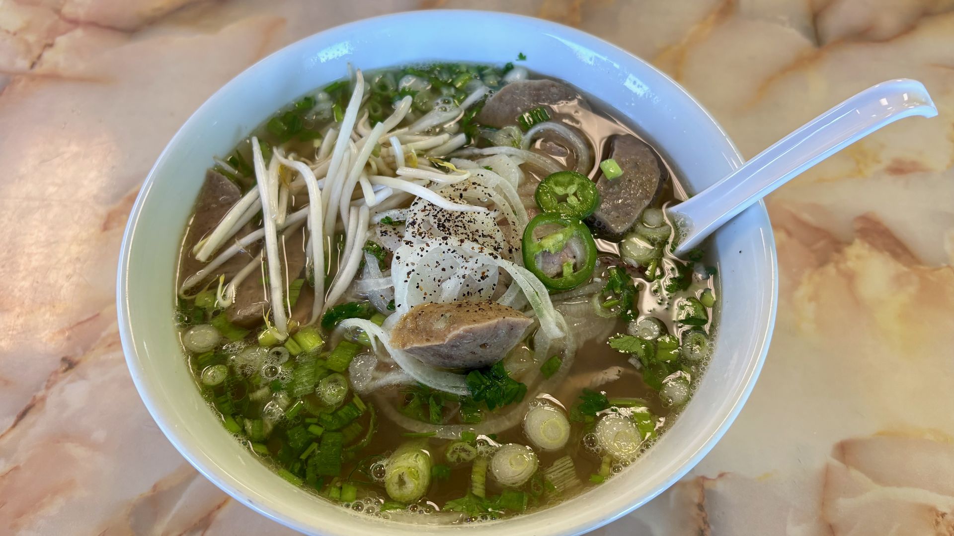 White bowl of Vietnamese pho with clear broth, meat slices, bean sprouts, onion, green herbs, sliced jalapeño, and a white soup spoon on a marble table.