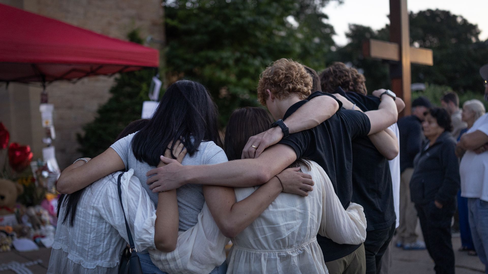 Back view of a line of people with arms around each other in a group hug outdoors. They wear casual clothing; a red canopy and brick wall on the left, and a wooden cross with trees in the background.