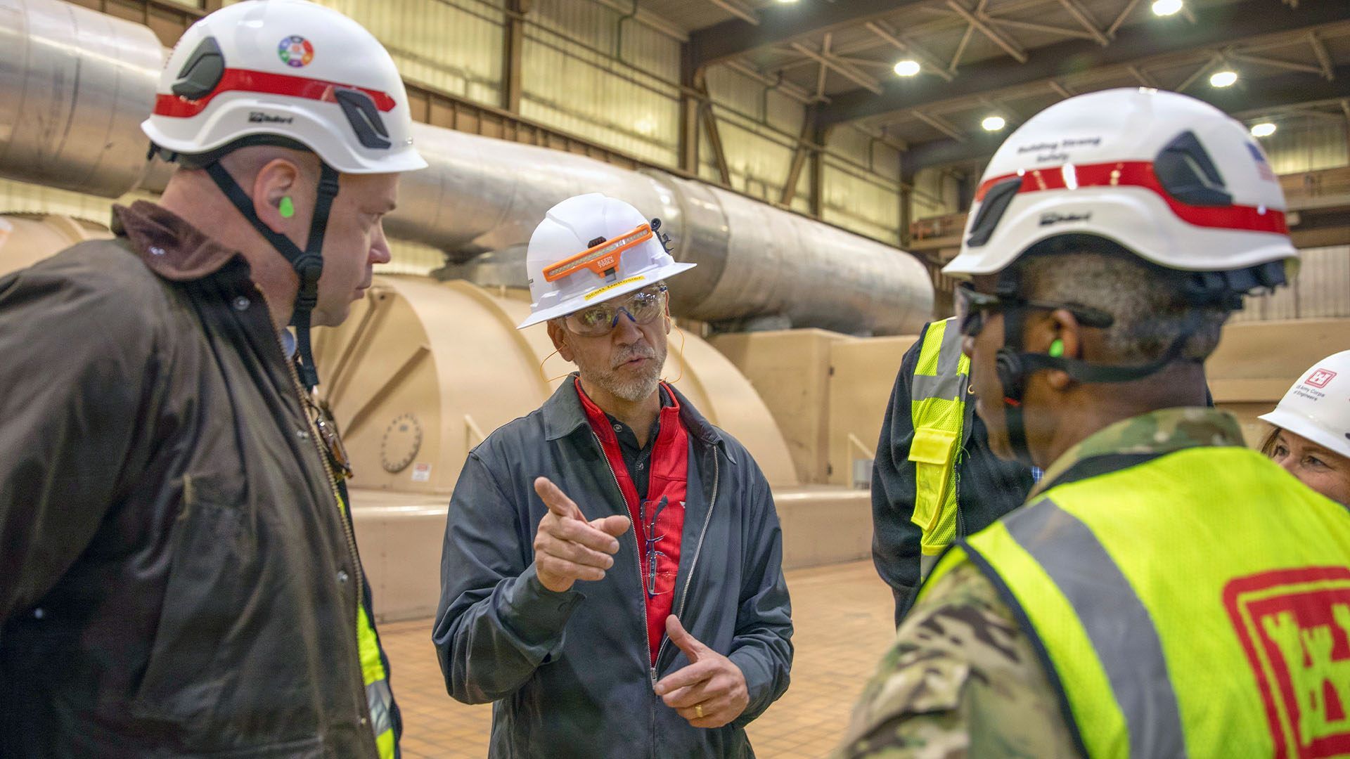 Group of factory workers in hard hats and high-visibility vests discuss near a large cylindrical turbine inside an industrial facility; the central man points while speaking.
