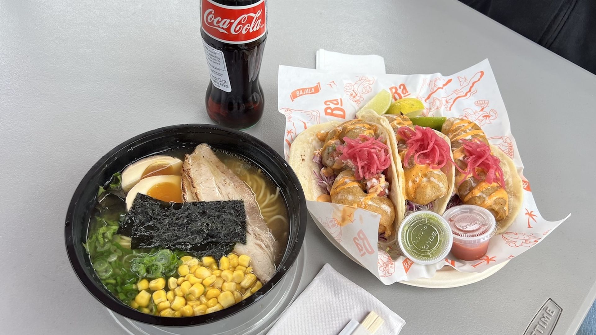 A photo of a bowl of ramen and a plate of three fish tacos, with a bottle of coca-cola in the background.