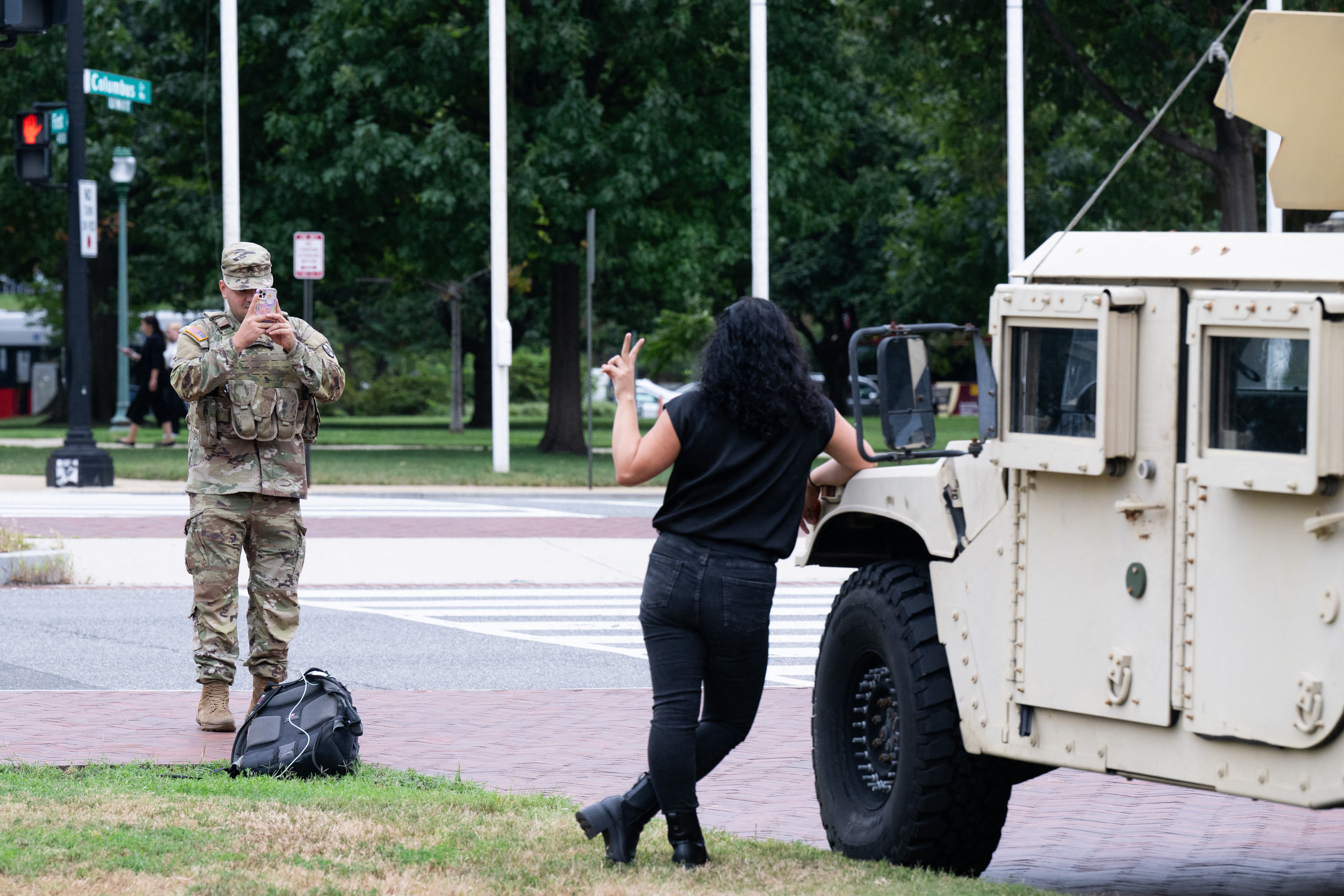 A soldier in camouflage uniform takes a photo with a smartphone of a woman posing with a peace sign next to a tan military vehicle on a street corner with trees in the background.