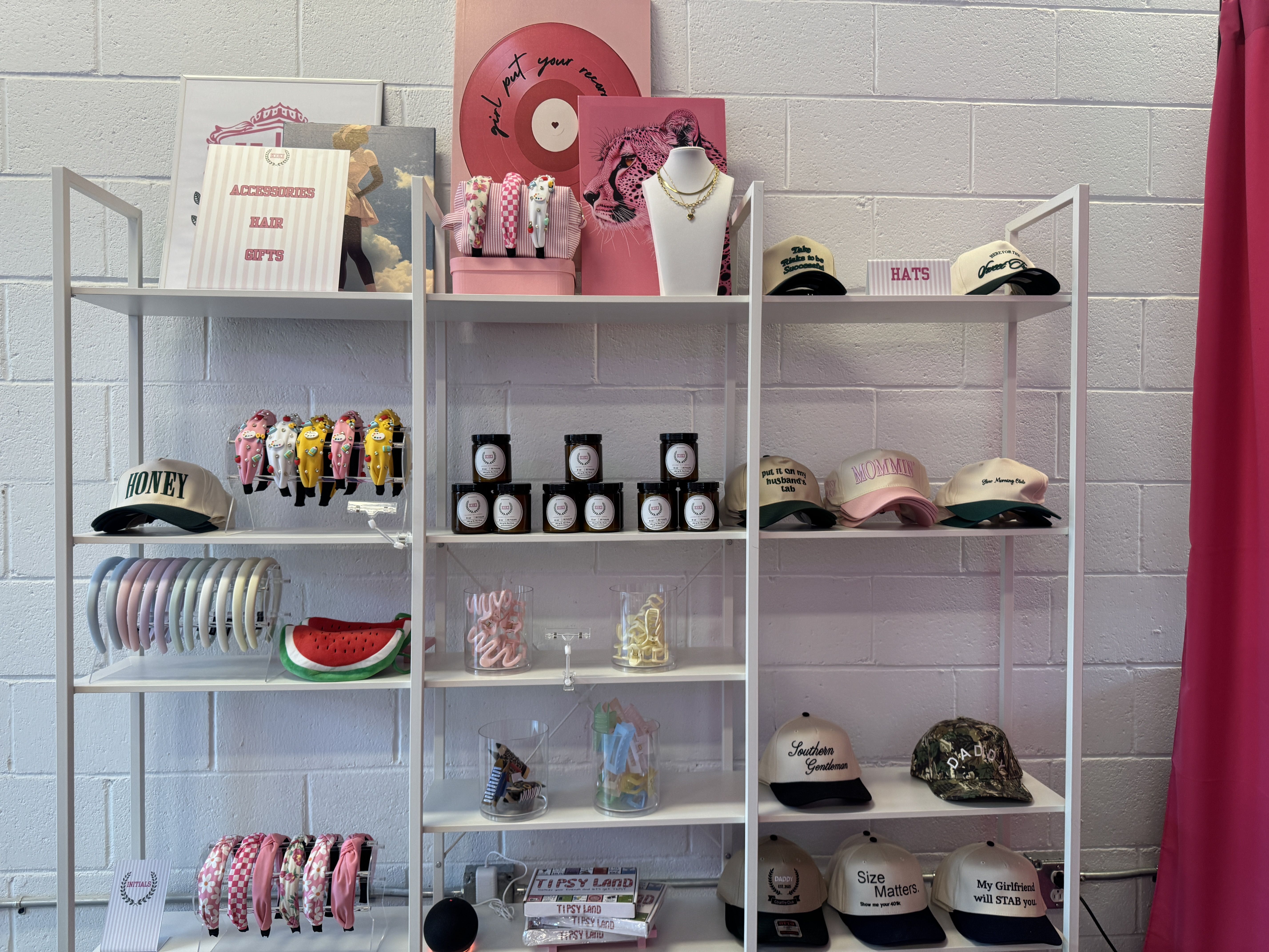 White metal shelves displaying pink, cream, and camouflage hats with various text, pink and white headbands, hair clips in clear containers, jars of product, and pink-themed decorative prints.