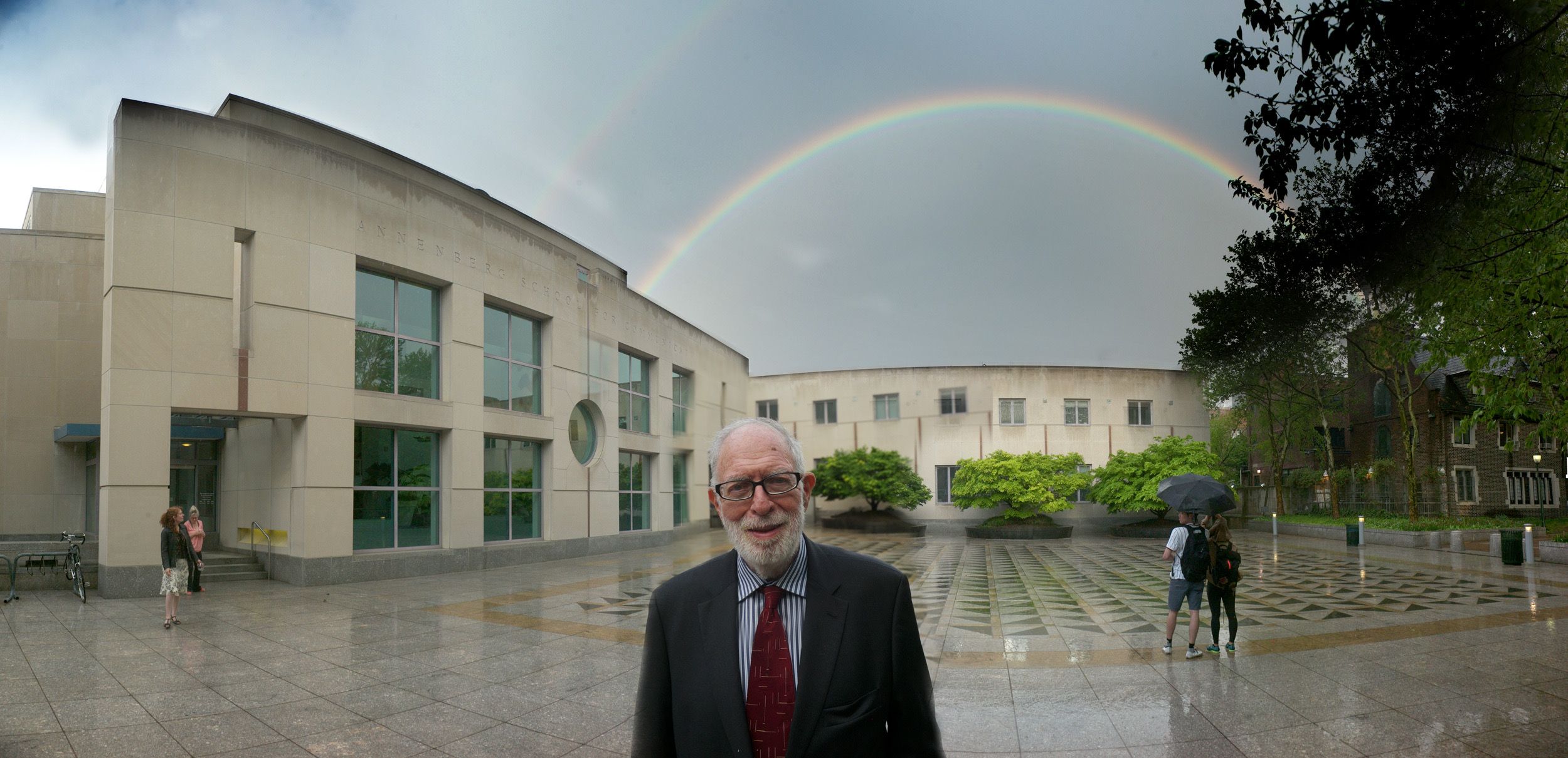 Panoramic view of a beige modern building with windows, a rainbow arching across a gray sky, and a man in a suit in the foreground. Wet pavement reflects the rainbow; trees on the right.