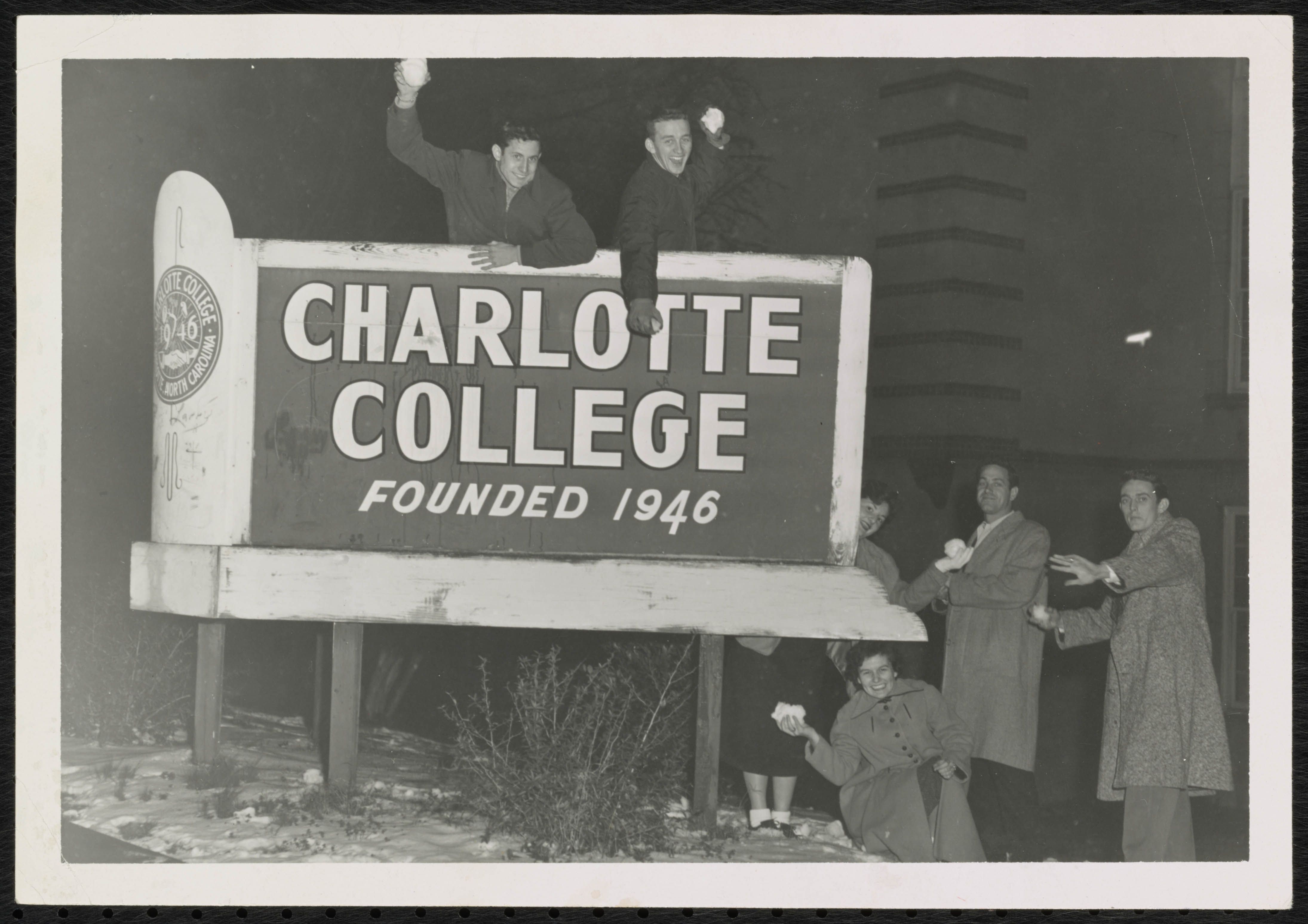 Black-and-white photo of several people atop a large sign that reads "CHARLOTTE COLLEGE" founded 1946; two men raise fists from the railing while others smile nearby.