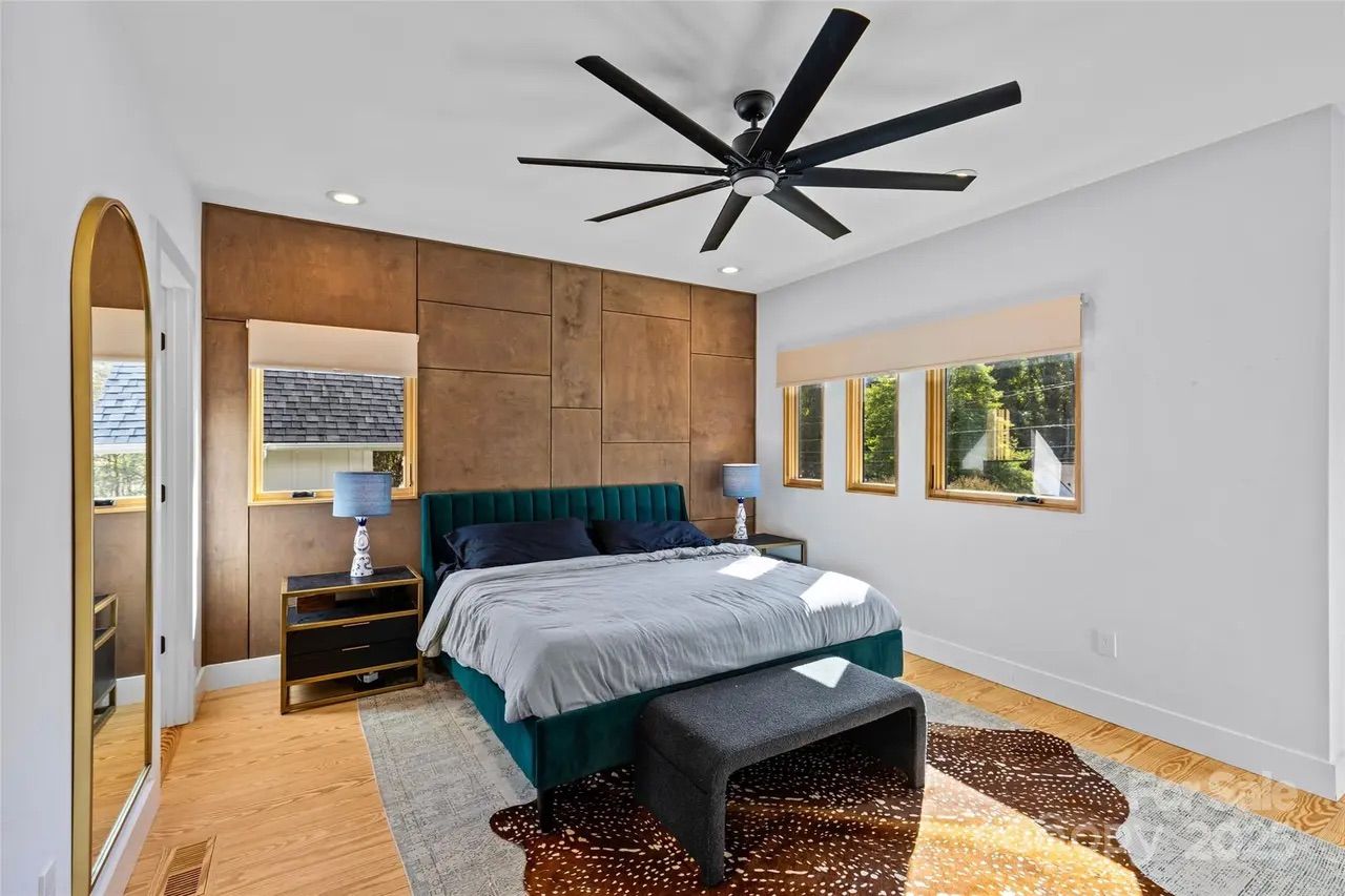 Bedroom with green upholstered bed, gray bedding, brown wooden accent wall, black ceiling fan, two blue lamps on black nightstands, three windows with beige shades, and a patterned rug.