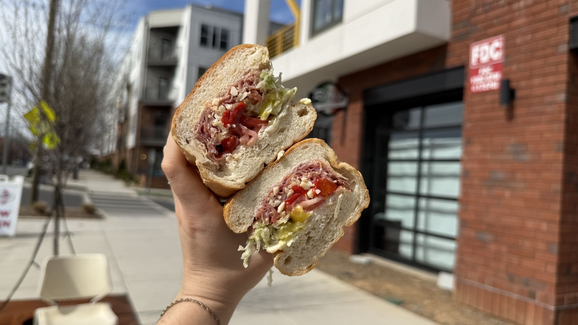 Hand holds a sliced baguette sandwich with ham, lettuce, tomato and cheese; outdoors on a sunny urban street with brick buildings and modern storefronts in the background.