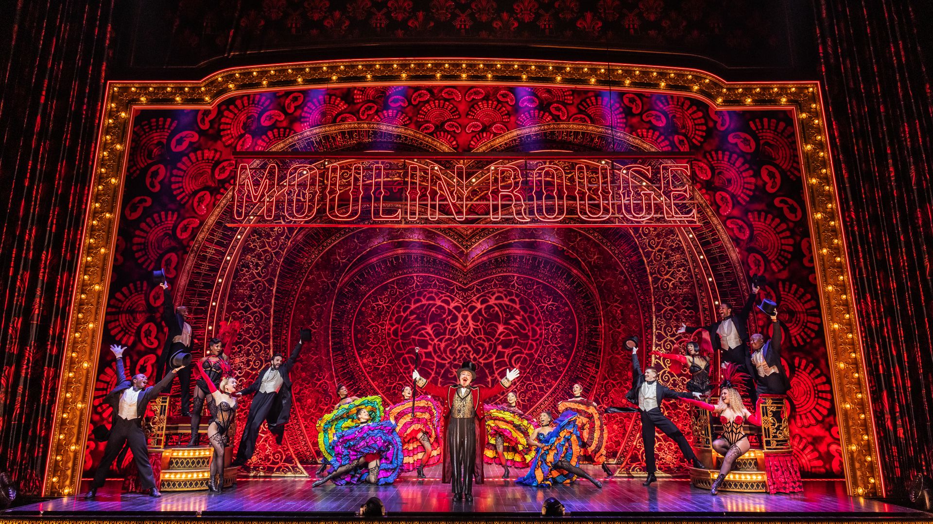 Onstage at "Moulin Rouge", a neon sign lights a gold-trimmed arch and ornate red backdrop as dancers in colorful costumes perform under warm stage lights.