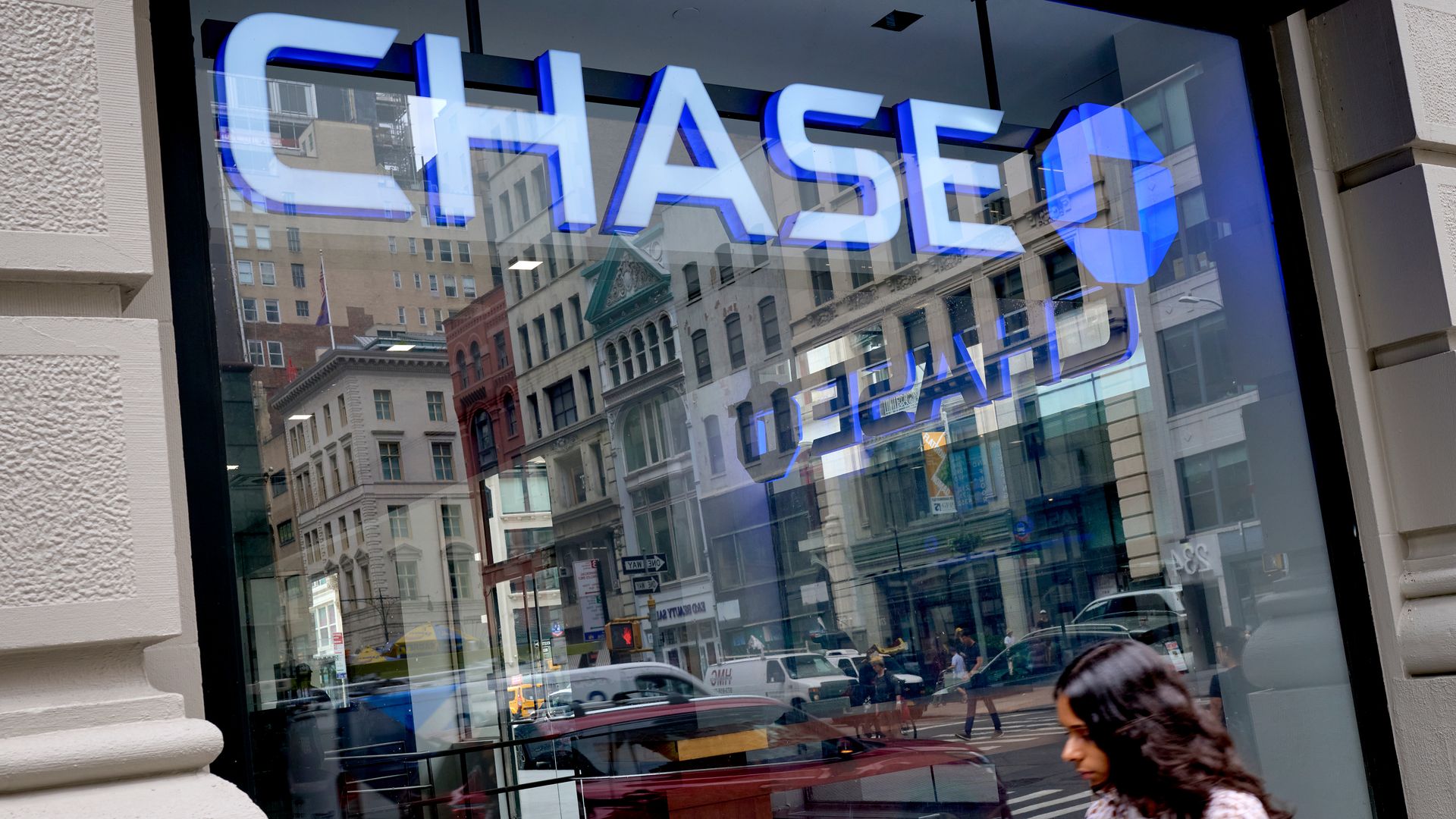 Storefront window with a large blue illuminated CHASE sign, reflections of city buildings on the glass, and a woman in a patterned dress walking by while holding a phone.