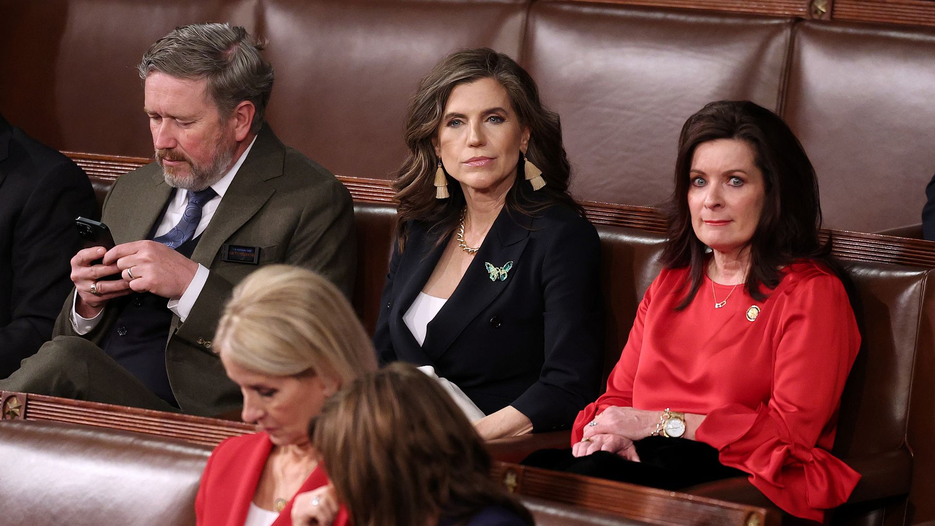Three individuals seated on brown leather benches; man in olive suit using phone, woman in navy blazer with butterfly brooch looking ahead, woman in red dress sitting with hands folded.