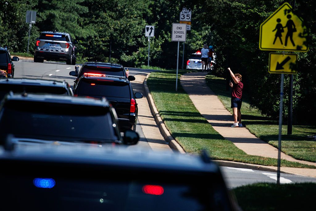 Five black SUVs and two state trooper vehicles drive up a road as a man on the sidewalk raises his fist in the air, watching them. Traffic signs show they are driving through a school zone with a 35 speed limit.