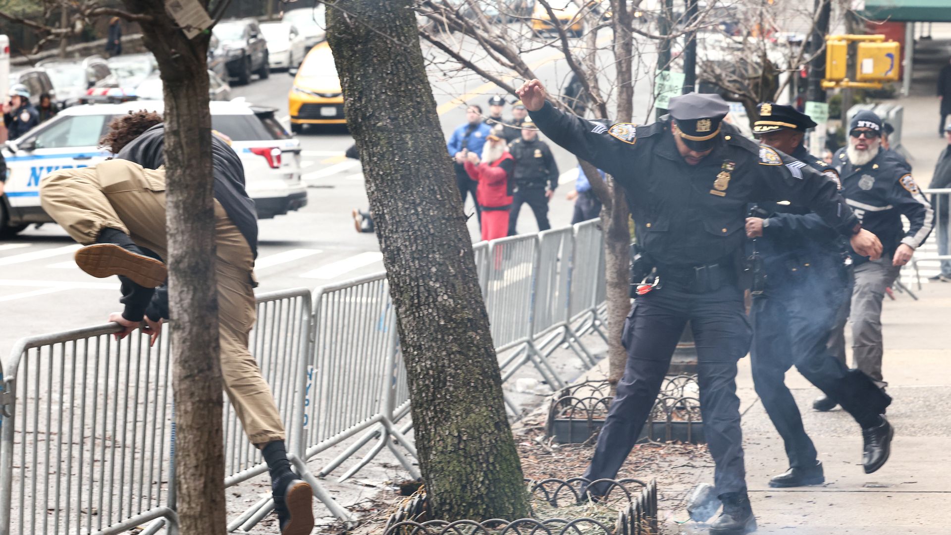 Person climbing over metal barricade on a city street while police officers react nearby, with some smoke visible on the sidewalk and a few bystanders in the background.