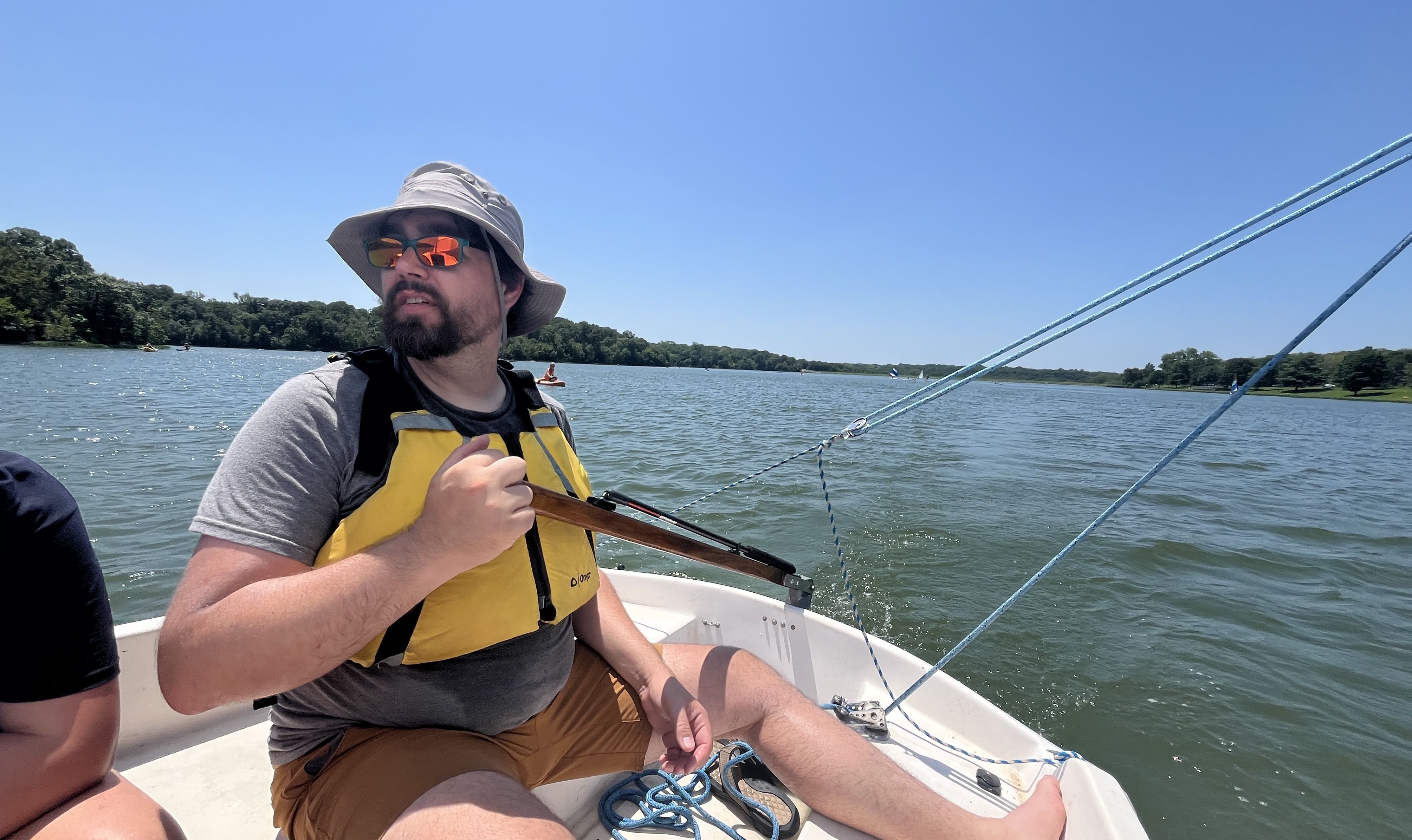 Man wearing a gray hat, sunglasses, yellow life jacket, and brown shorts sailing a small boat on a sunny lake with trees and clear blue sky in the background.
