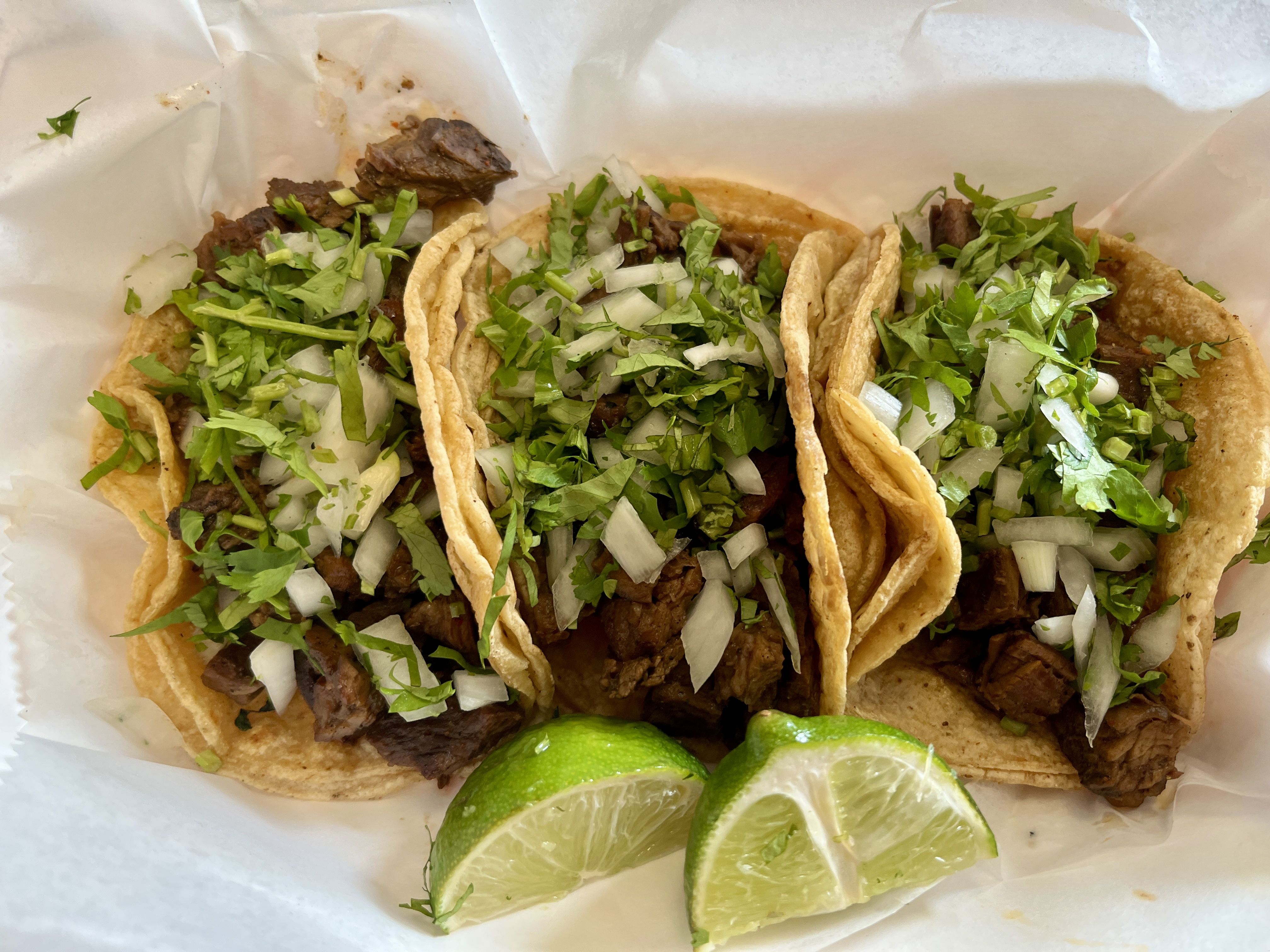 Three carne asada tacos with chopped onions and cilantro in corn tortillas, served with two lime wedges on white paper.